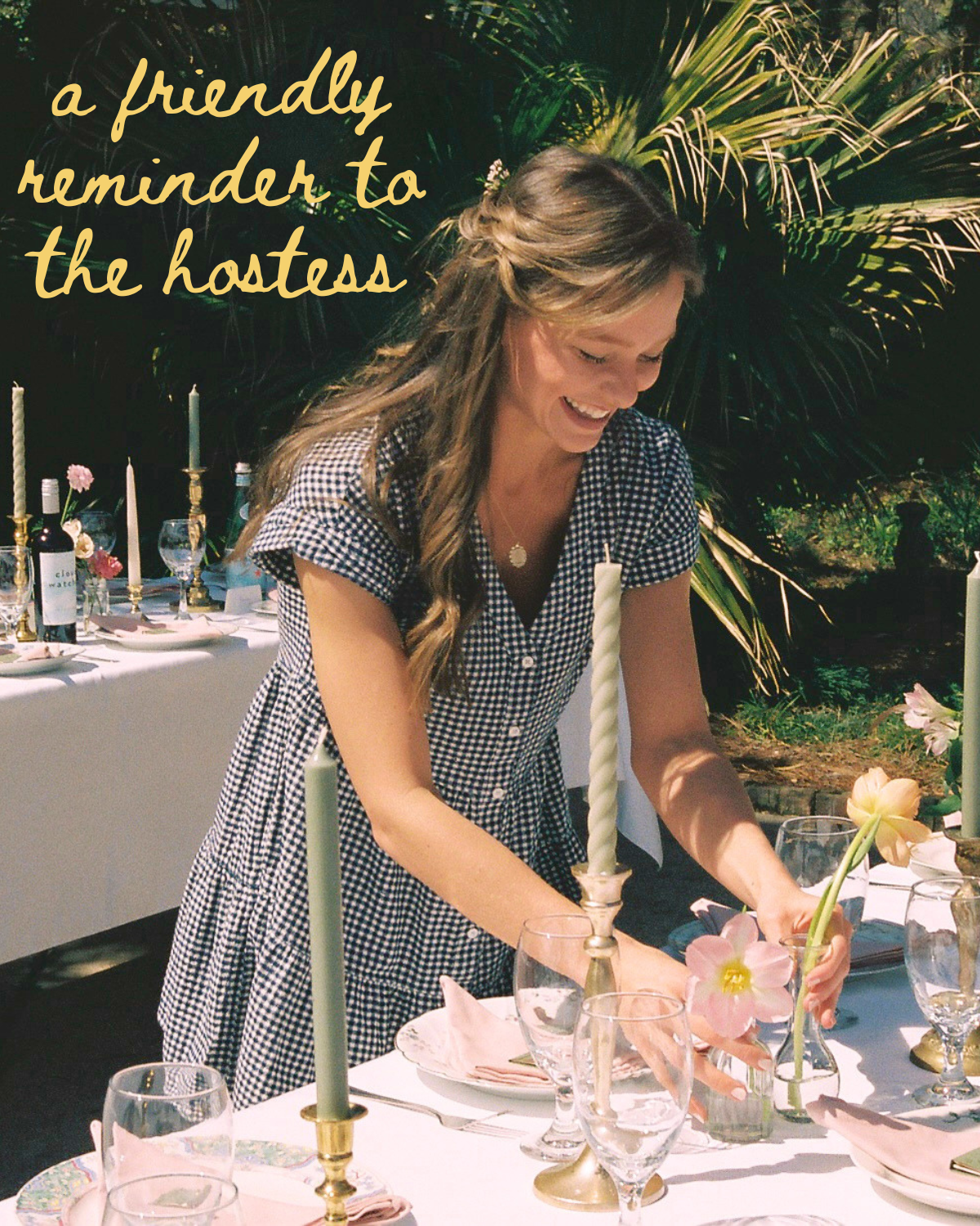 A woman in a gingham dress smiles while arranging flowers on an outdoor dining table decorated with candles, glassware, and pink flowers. Green foliage is in the background.