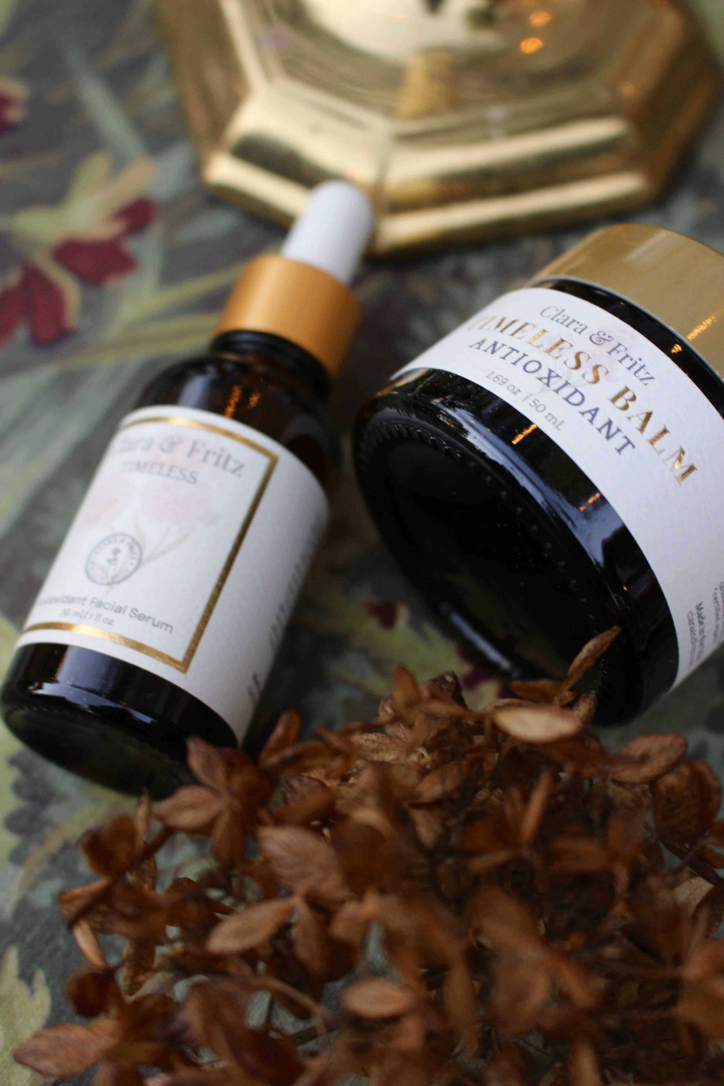 Beauty serum bottles, one with a dropper and the other labeled 'LAVIELES BALM ANTIOXIDANT', placed on a floral patterned surface with dried flowers and a decorative gold-edged mirror.