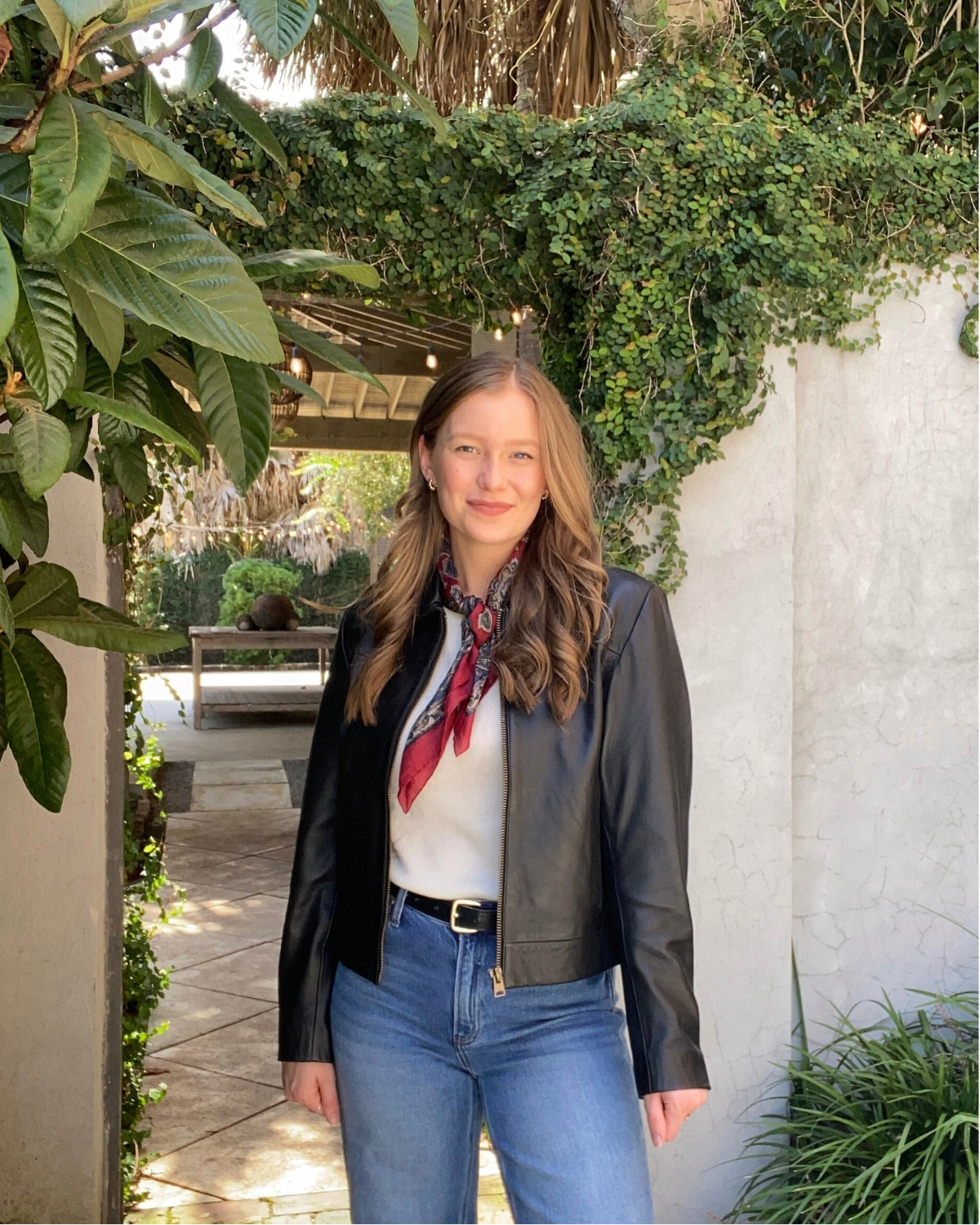 A woman with light brown hair and fair skin standing outdoors in a garden area with lush green foliage and a white wall. She is wearing a black leather jacket, a light-colored top, blue jeans, and a red patterned scarf around her neck.