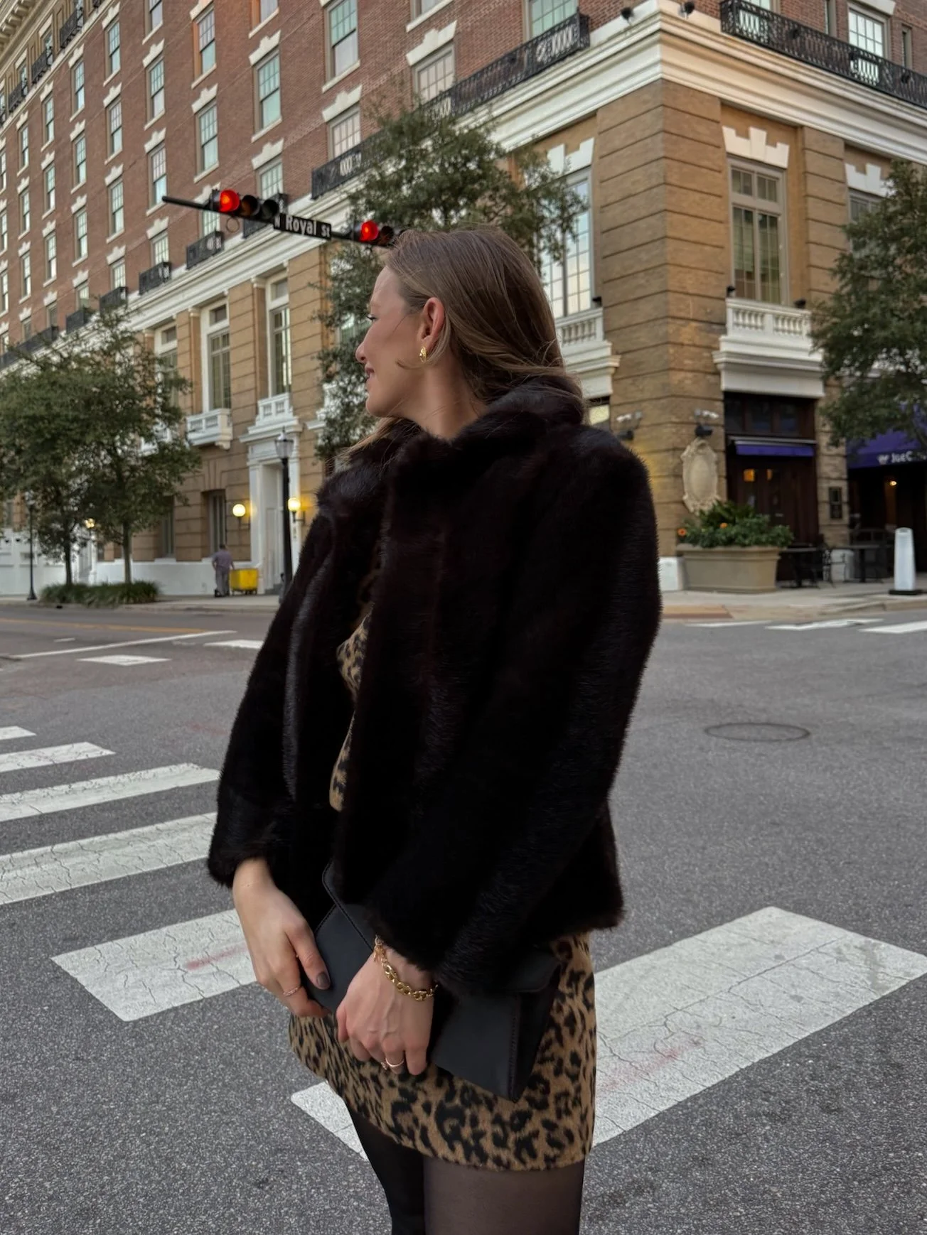 A woman with shoulder-length brown hair, wearing a black fur coat, leopard print dress, and holding a black clutch, stands on a city street corner at dusk, with a building, trees, and a traffic light in the background.