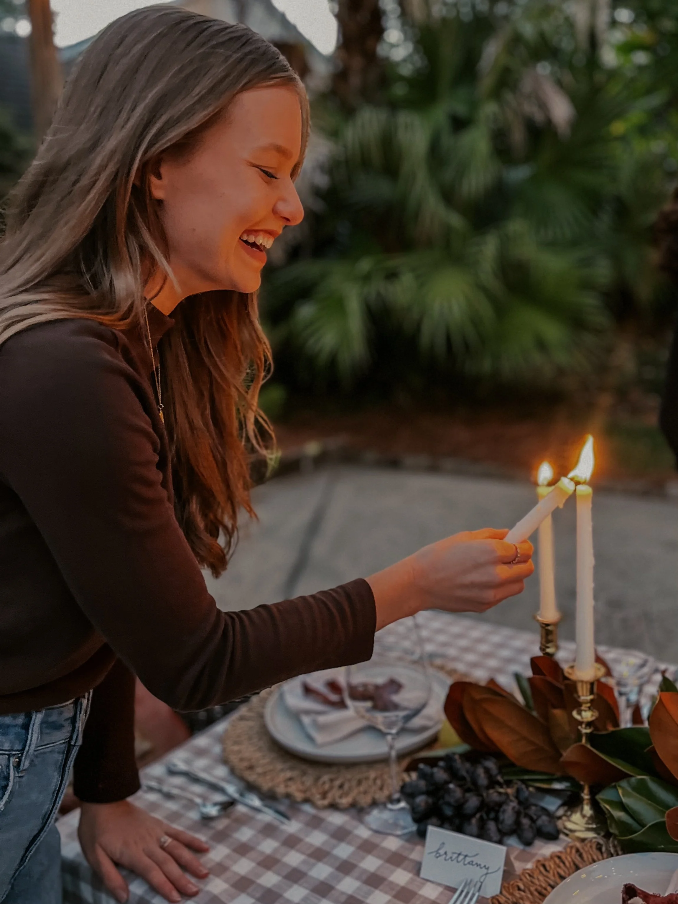 A woman lighting candles on a table set for a meal, with a brown checkered tablecloth, plates, napkins, and grapes as decor, outdoors with greenery in the background.