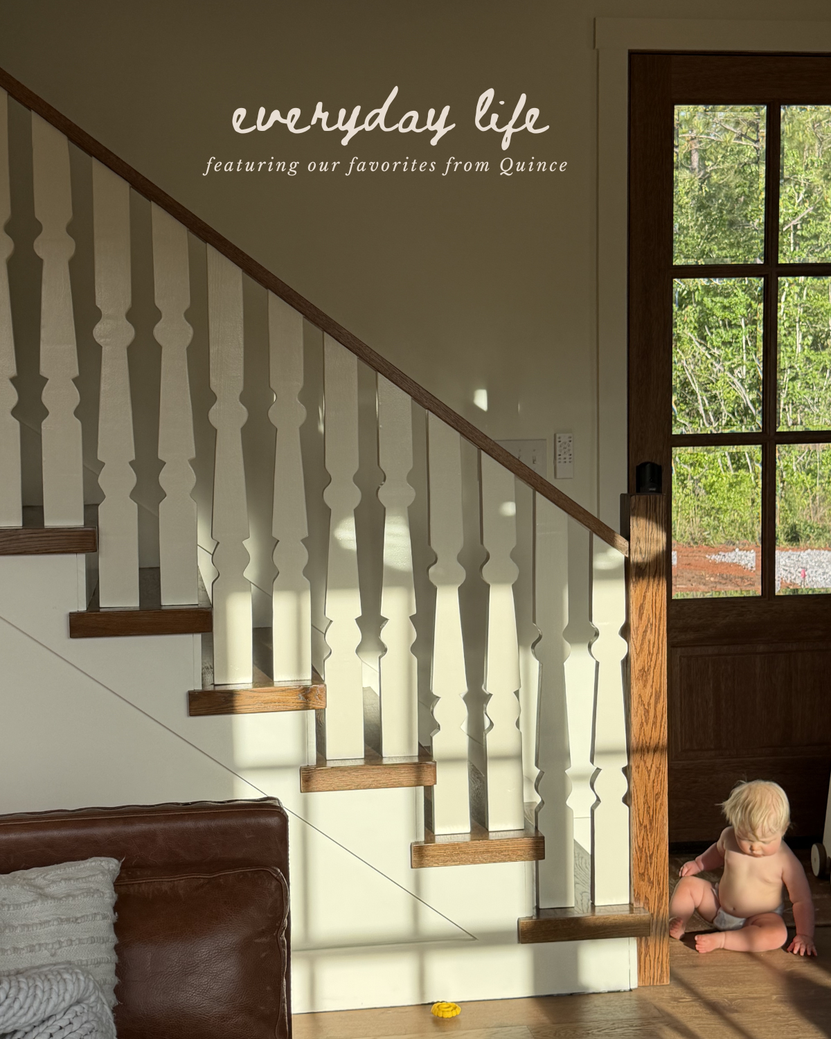A staircase with white spindles and wooden handrails, sunlight casting shadows on the wall, a large window showing greenery outside, and a baby sitting on the floor near the door.