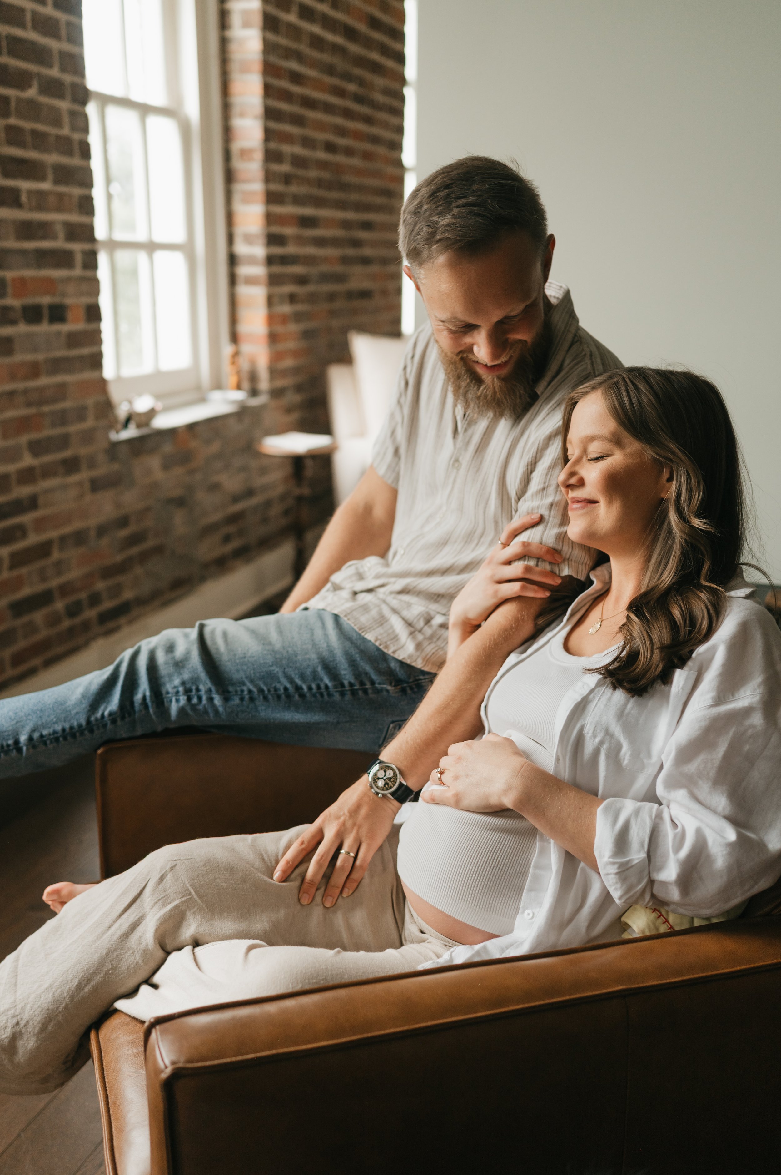 A smiling man and pregnant woman sitting on a leather couch in a cozy, modern living room illuminated by natural light.