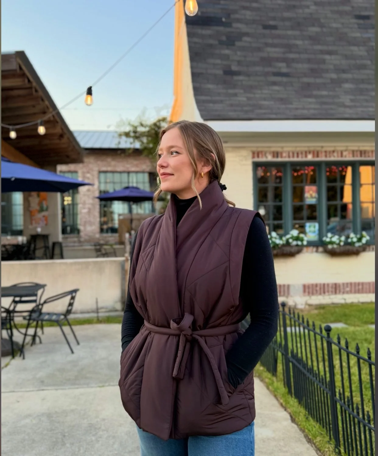 A woman standing outdoors in front of a restaurant, wearing a dark vest over a black long-sleeve shirt, with a black hair tie, and jewelry earrings, with a relaxed expression.