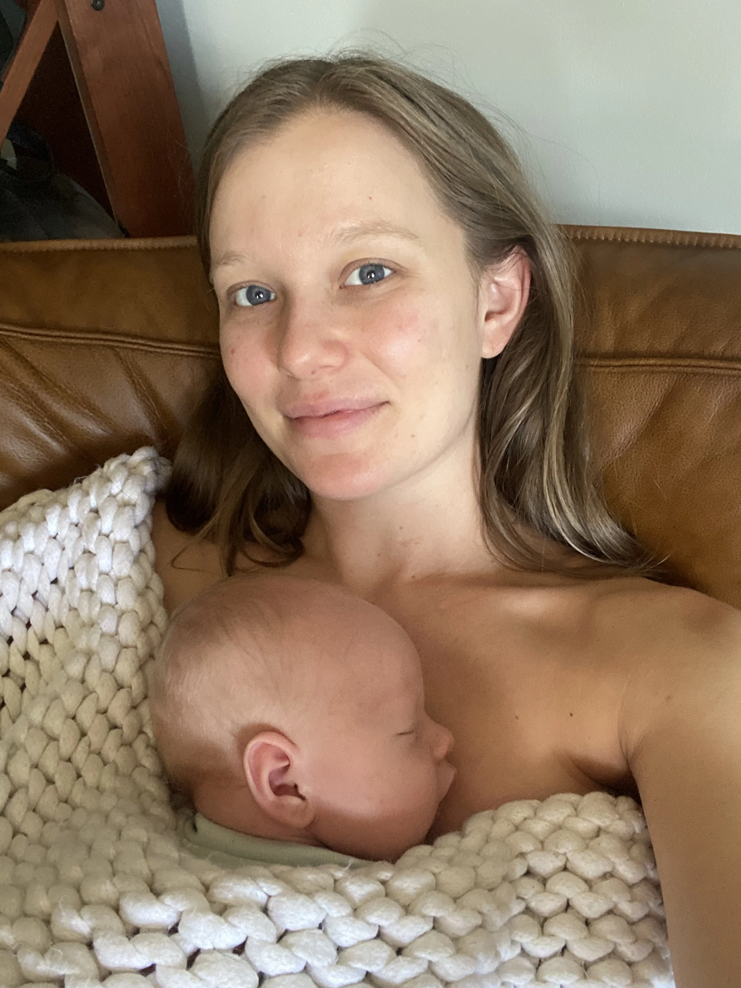 A woman with light skin and brown hair holding a sleeping baby with light skin and blonde hair, both resting on a chunky cream-colored knit blanket on a brown leather couch.