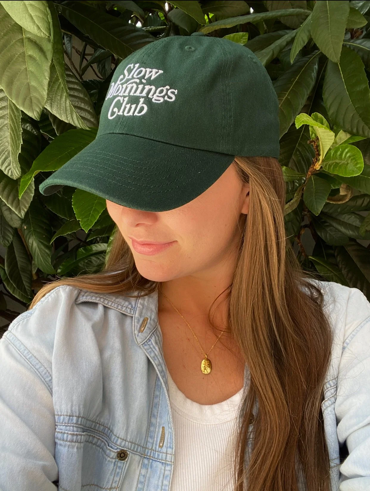 Woman wearing a green "Slow Mornings Club" cap, light denim jacket, and necklace, with green leaves in the background.