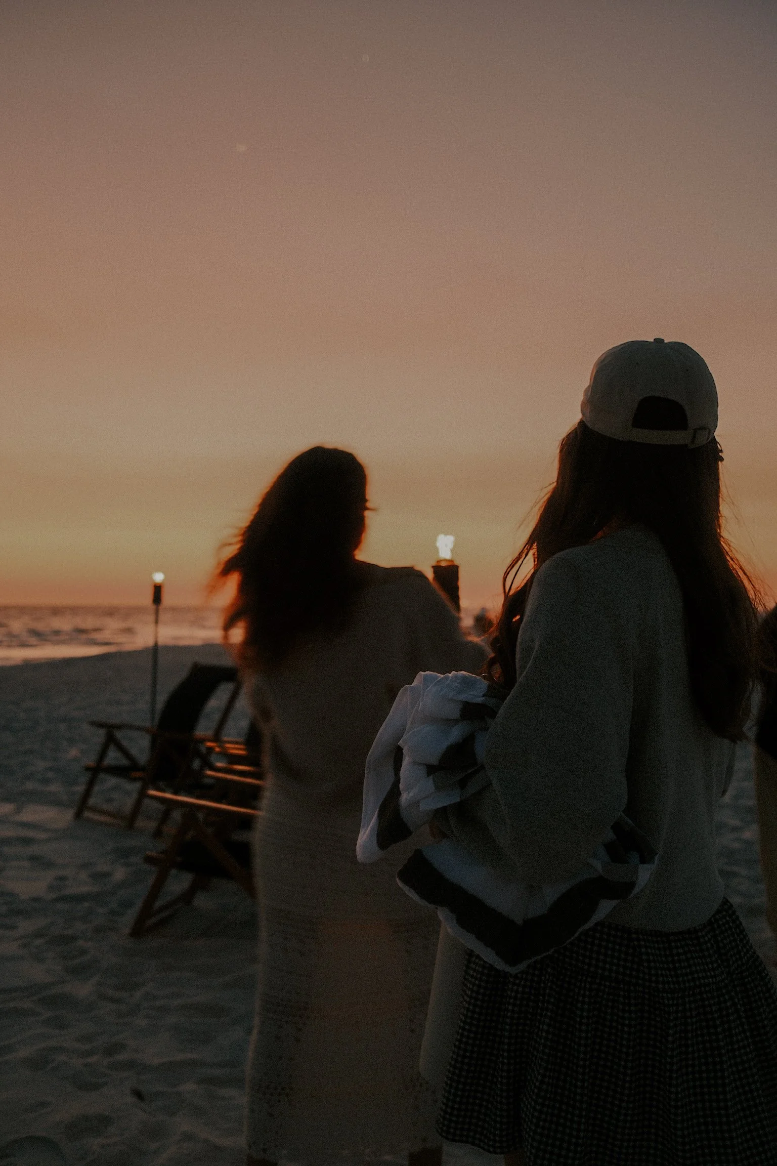 Two people at a beach during sunset, one holding a towel, with lounge chairs and tiki torches in the background.