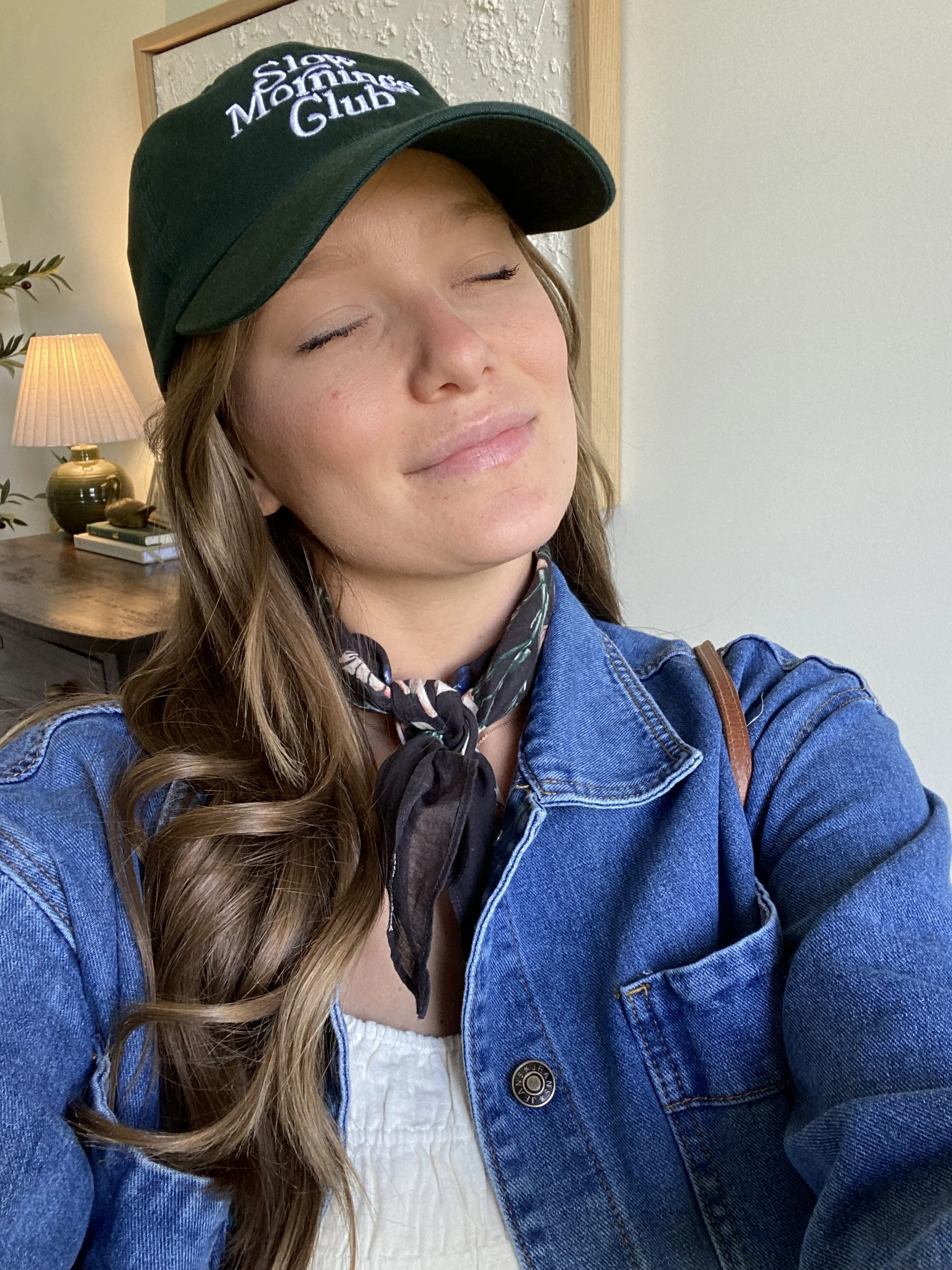 A woman with wavy brown hair, wearing a green baseball cap with white embroidered text, a denim jacket, a black scarf, and a white top, smiling with her eyes closed indoors near a table with a lamp and a plant in the background.