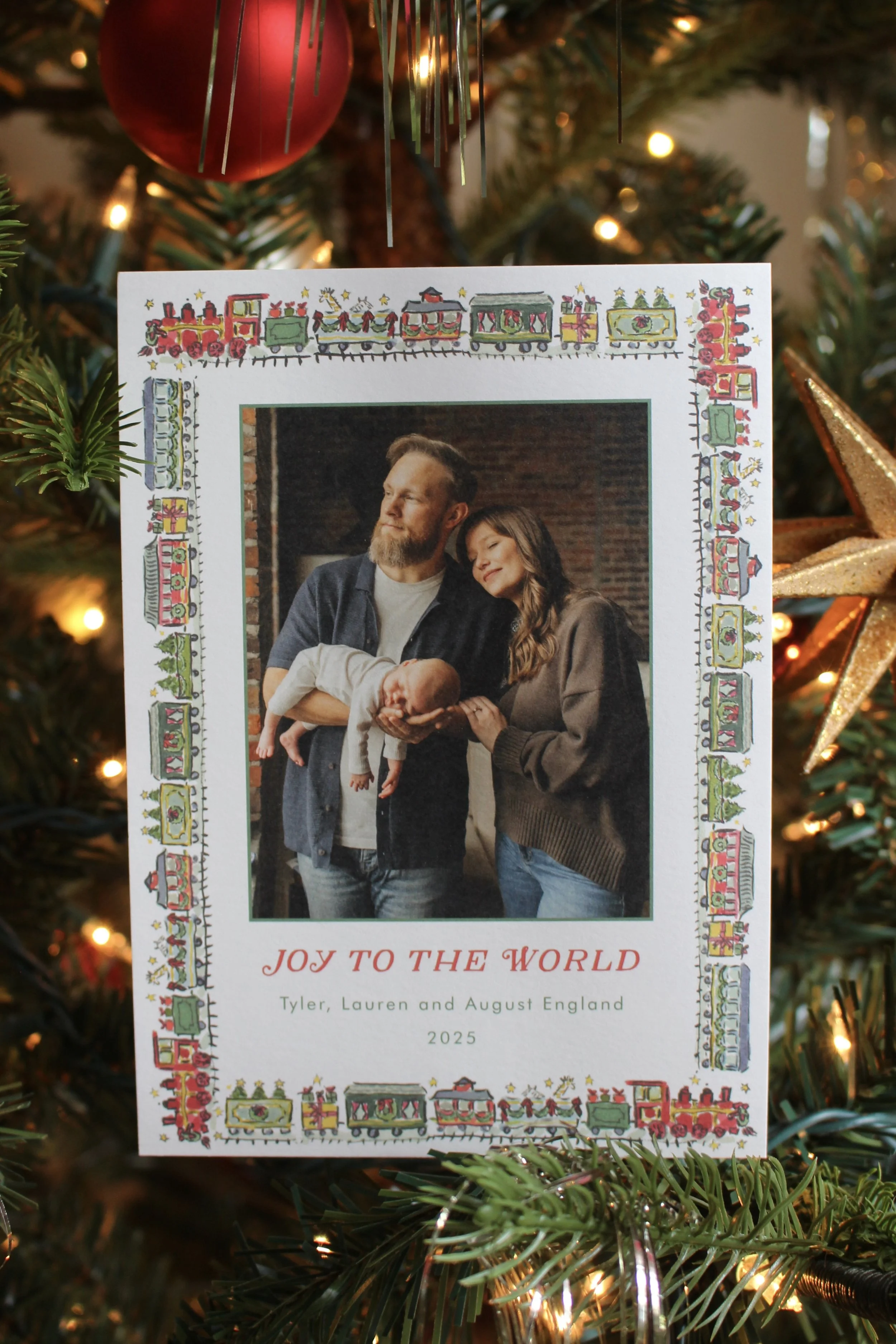 A holiday greeting card hanging on a decorated Christmas tree, featuring a photograph of a couple with their newborn, surrounded by colorful Christmas-themed illustrations and the text 'Joy to the World, Tyler, Lauren and August England, 2025.'
