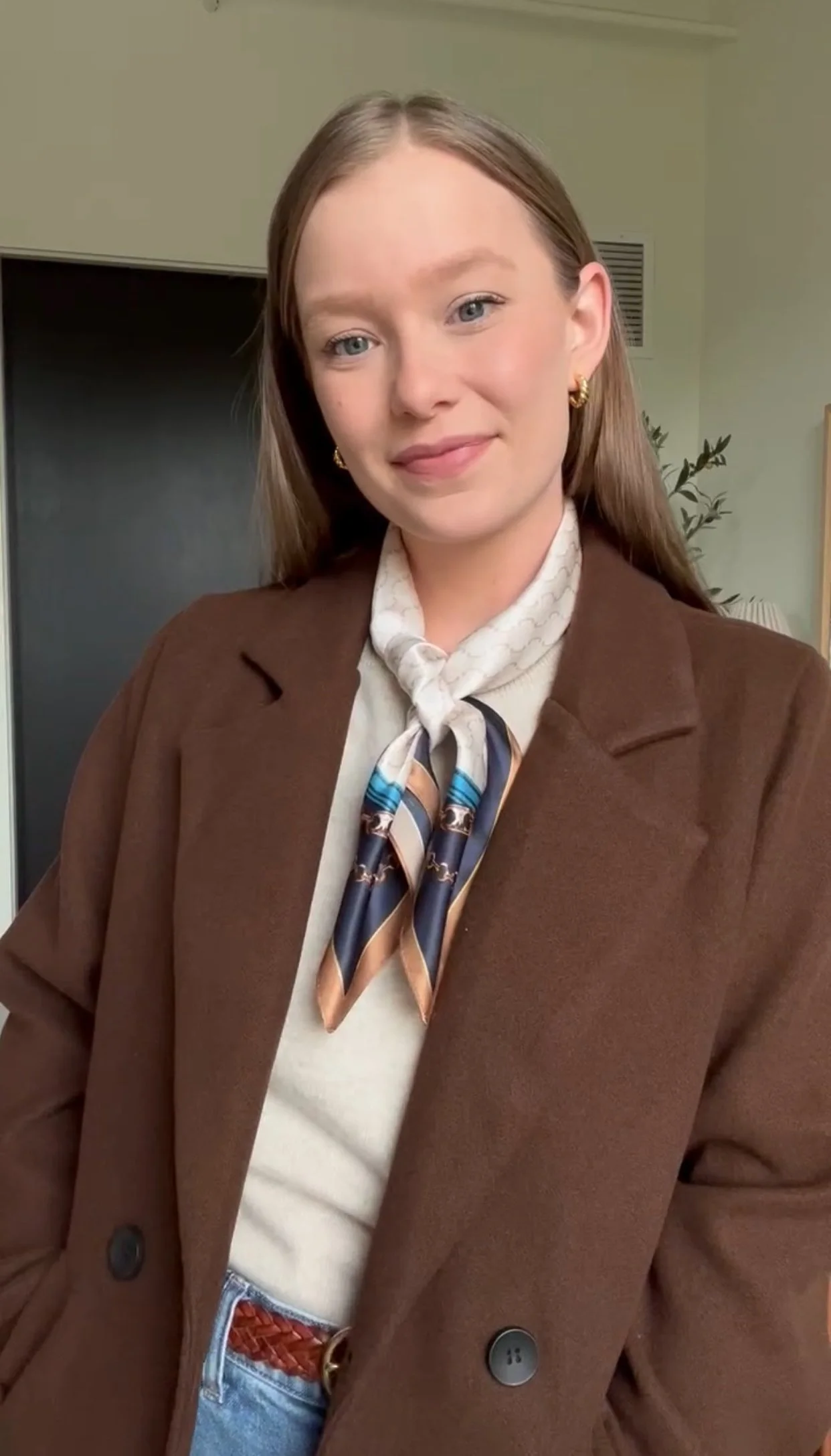 A young woman with light skin and long, straight brown hair smiling at the camera, wearing a brown blazer, a silk scarf, and gold earrings.