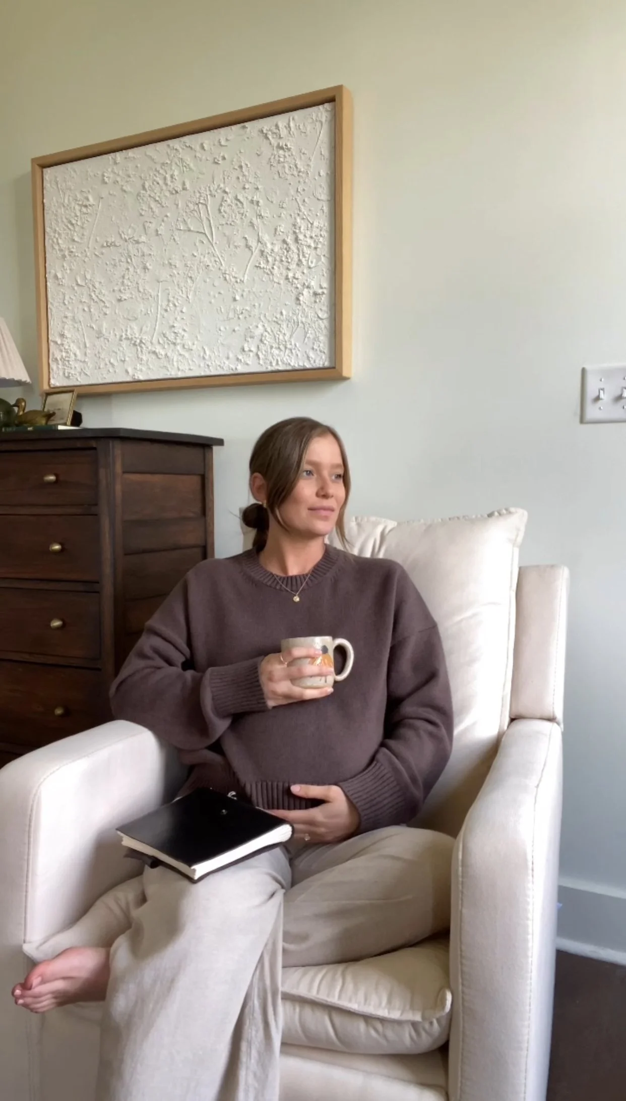 A woman with brown hair tied back, wearing a brown sweater and beige pants, sitting in a cream-colored armchair in a cozy living room. She is holding a ceramic mug in her right hand and a black notebook in her lap. Behind her, there is a dark wooden 