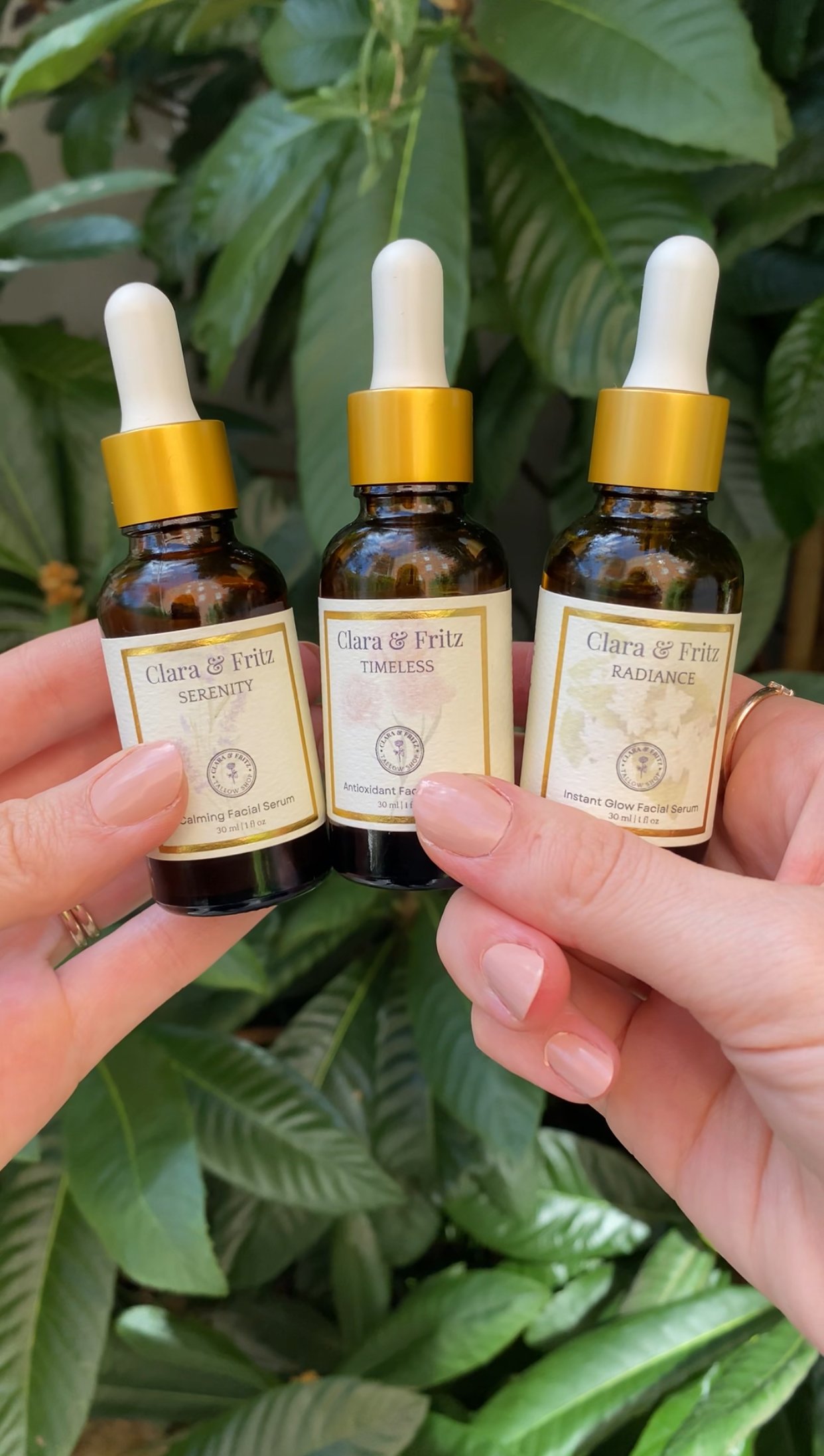 Three small brown glass bottles with golden dropper caps, labeled 'Clara & Fritz' with different names: Serenity, Timeless, Radiance. The bottles are held in two hands against a backdrop of green leafy plants.