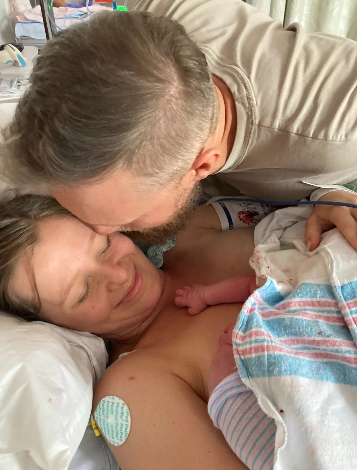 A woman in labor on a hospital bed holding a newborn baby, with a man leaning over and kissing her on the forehead.