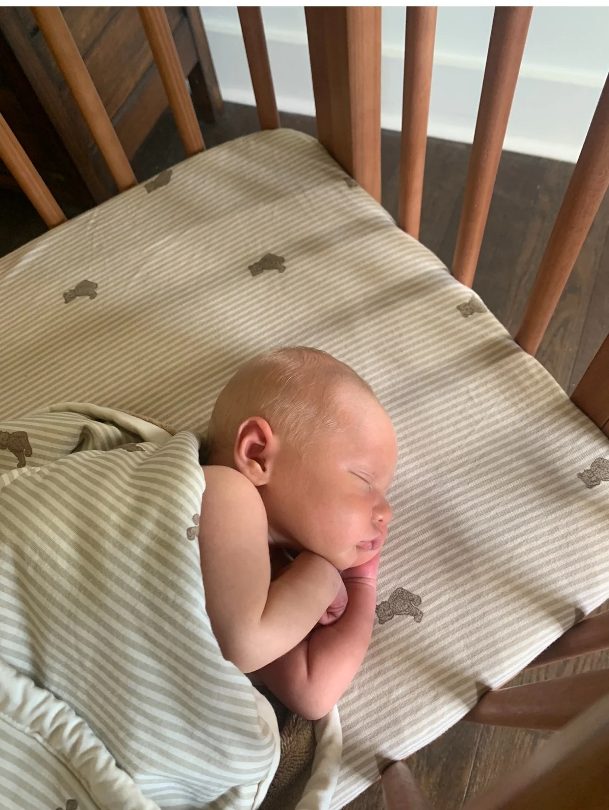 A newborn baby sleeping on a wooden crib mattress covered with a beige and white striped sheet with small brown teddy bear prints.