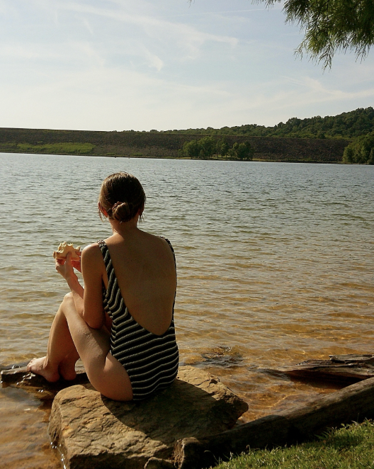 A woman sitting on a rock by a lake, eating a snack, with a forested area in the background.
