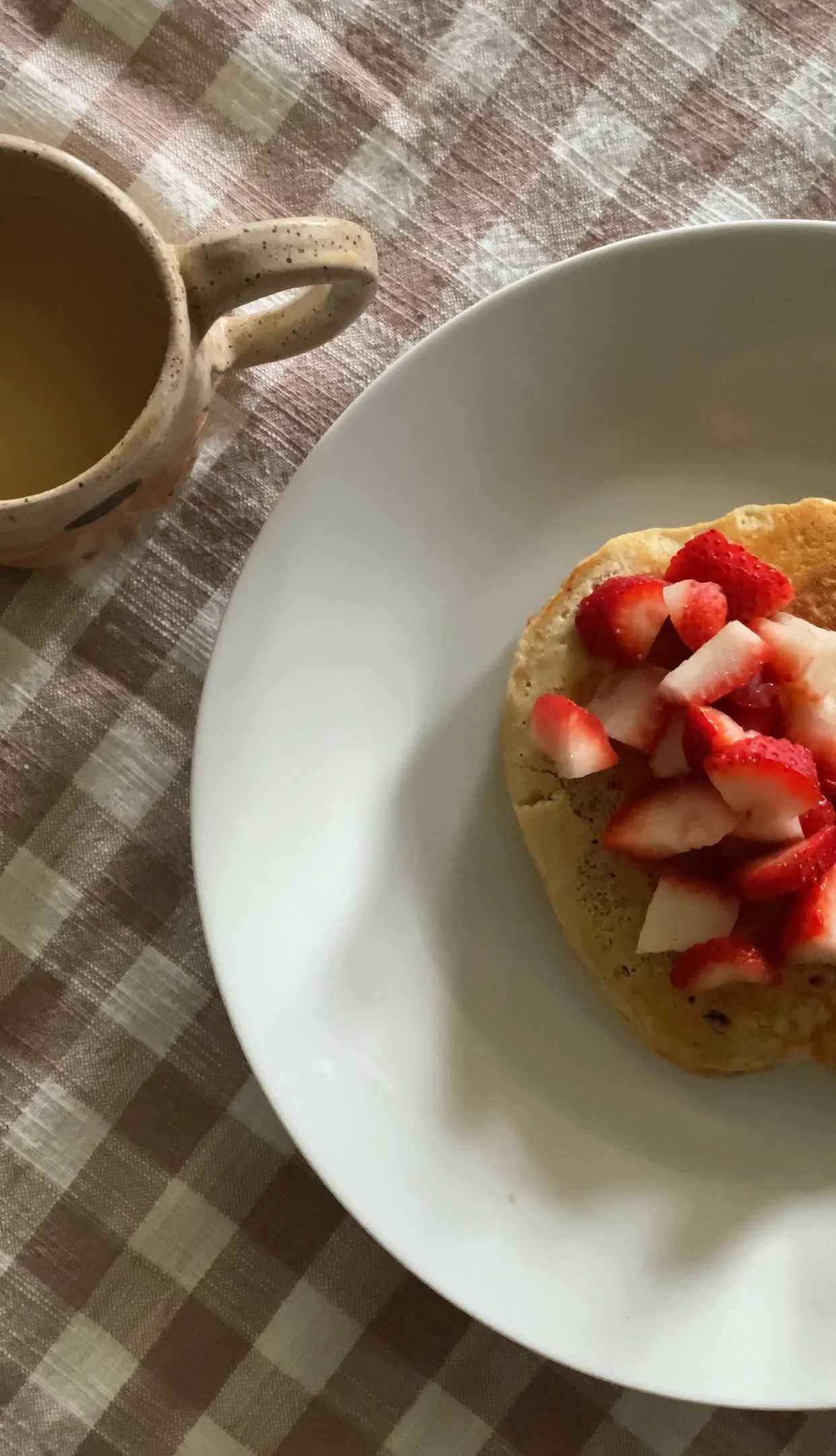 A plate with a dessert topped with sliced strawberries, next to a mug of coffee on a checkered tablecloth.
