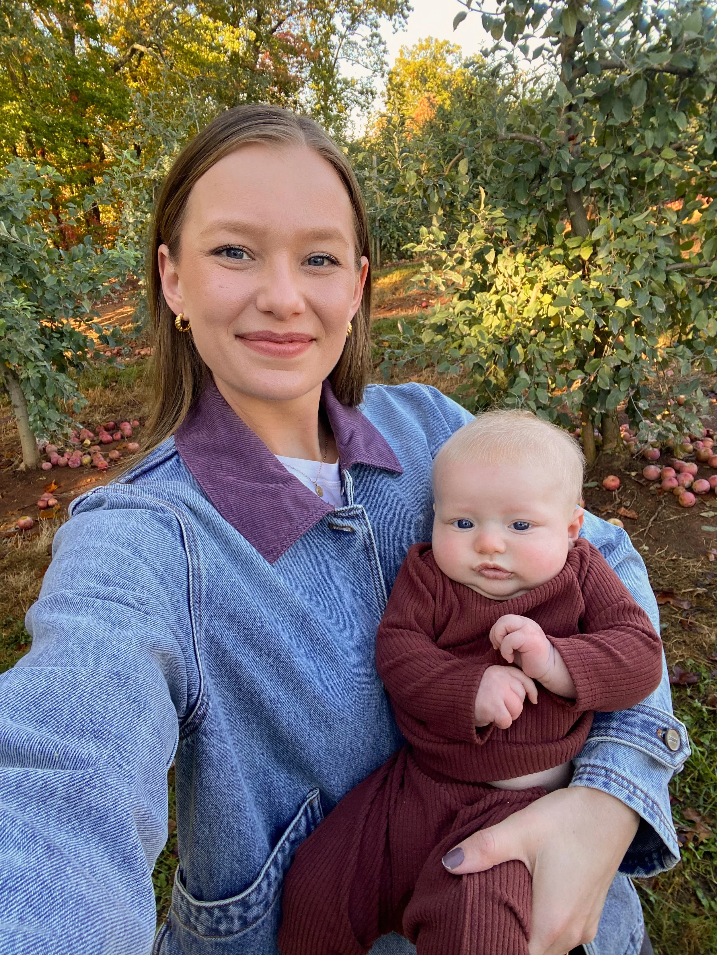 A woman with shoulder-length brown hair, blue eyes, wearing a denim jacket with a purple collar, is holding a baby with light blonde hair, blue eyes, dressed in a brown outfit. They are outdoors in an orchard with trees and fallen apples, during autu