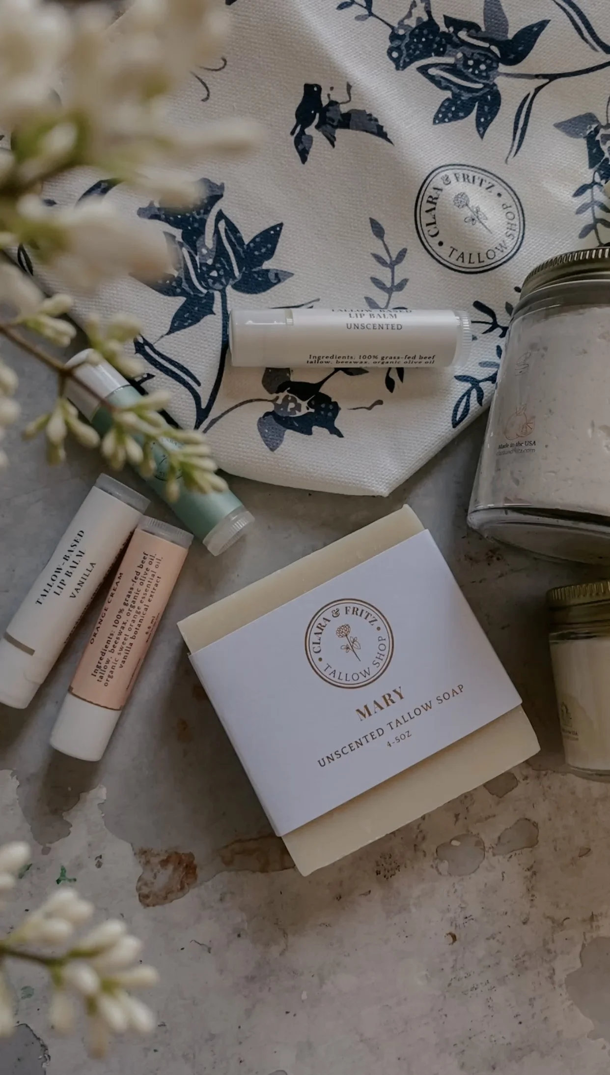Assorted tallow-based skincare products including unscented tallow soap, lip balm tubes, a jar of cream, and a cloth with floral patterns.