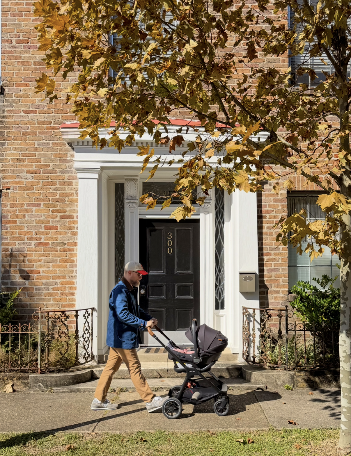 A person wearing a red and gray cap, dark blue jacket, and tan pants is walking on a sidewalk while pushing a black stroller in front of a brick house with a black door and white decorative trim. There is a tree with autumn leaves in front of the hou