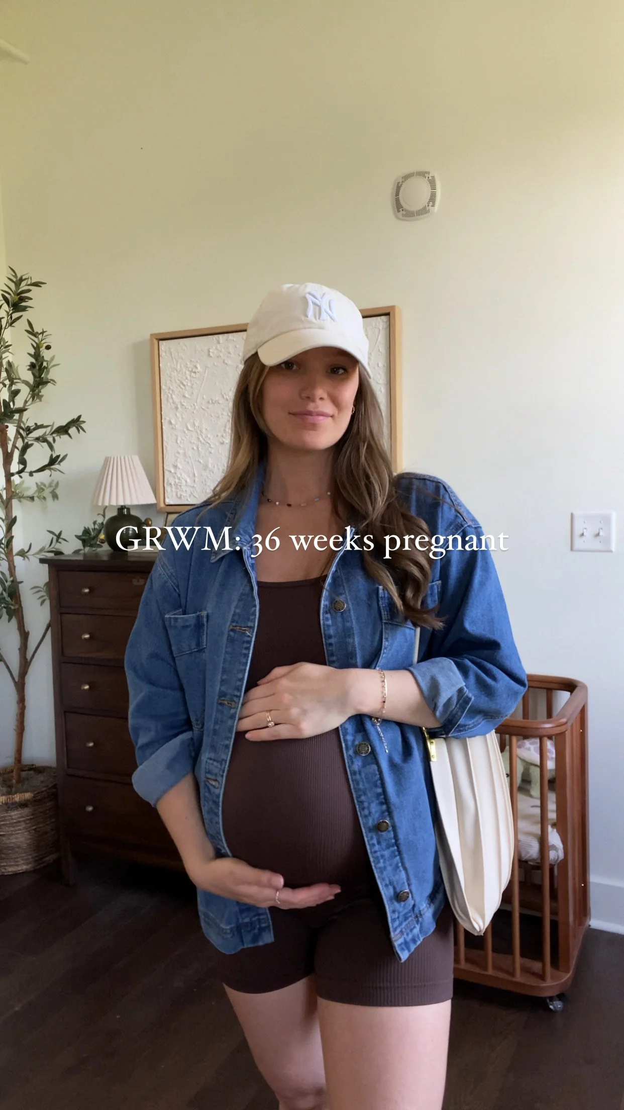 Pregnant woman with long hair wearing a denim jacket, black shorts, a white baseball cap, and jewelry, standing in a room with a wooden dresser and a potted plant, with text overlay indicating she is 36 weeks pregnant.
