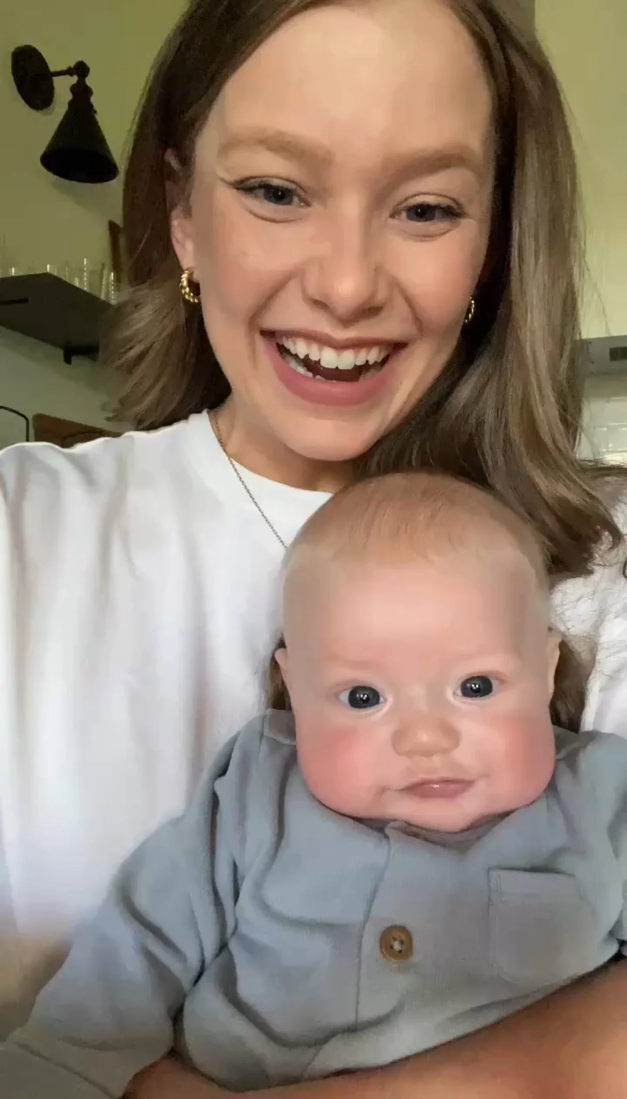 A woman with brown hair smiling while holding a baby with big blue eyes and a round face