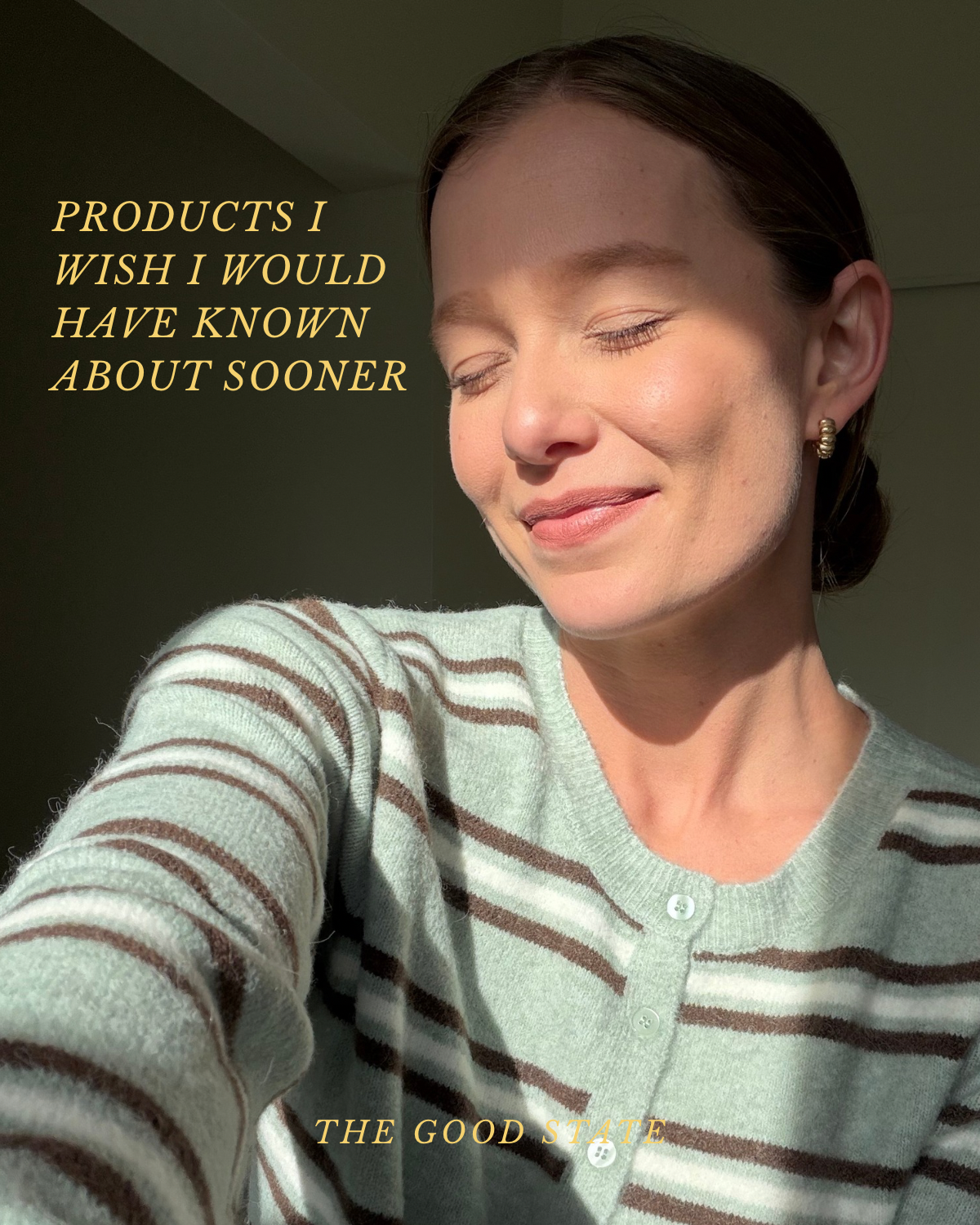 A woman with closed eyes smiling softly, wearing a light-colored sweater with brown stripes and gold earrings, bathed in sunlight in a softly lit room.