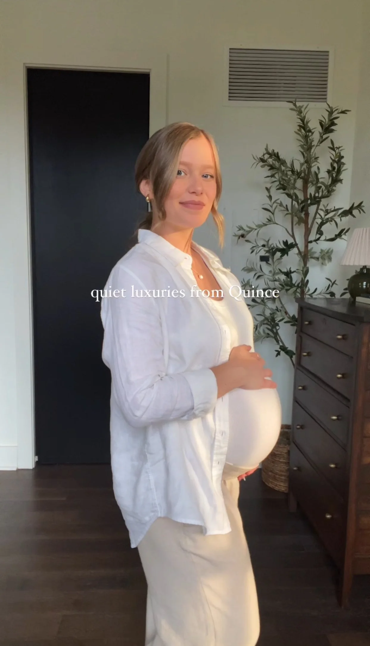A pregnant woman with blonde hair, wearing a white shirt and light-colored skirt, standing indoors in front of a dark door and a dresser with a lamp, with the text 'quiet luxuries from Quince' overlaid.