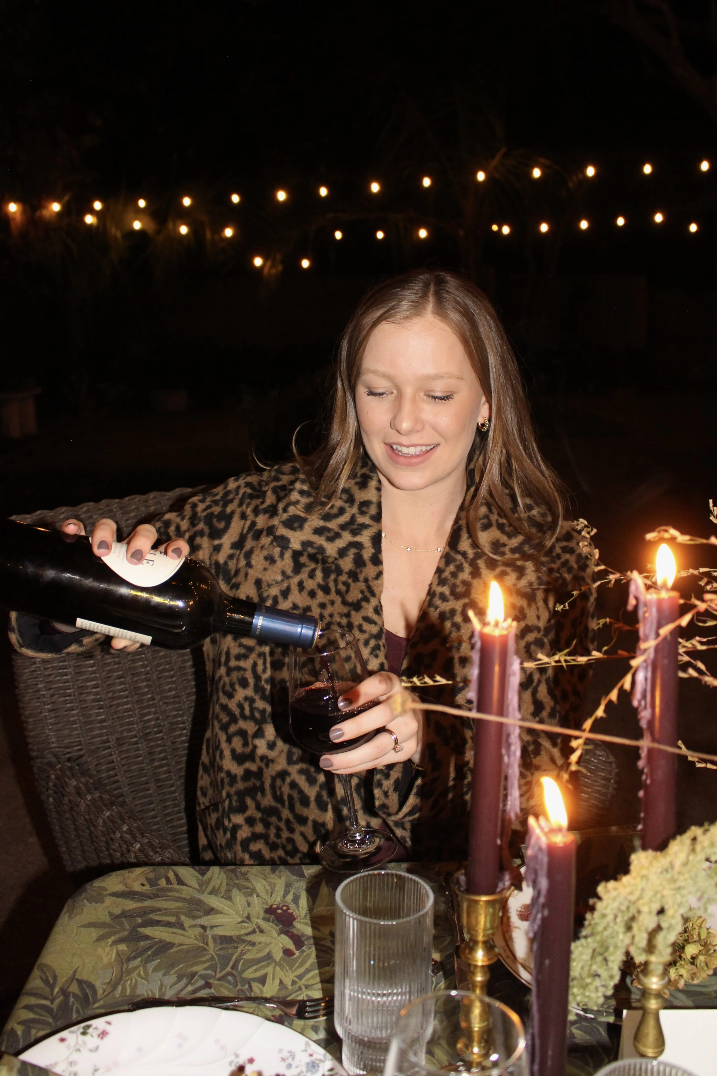 A woman with reddish brown hair wearing a leopard print coat is pouring red wine into a glass at a dinner table. There are candles and string lights in the background at night.