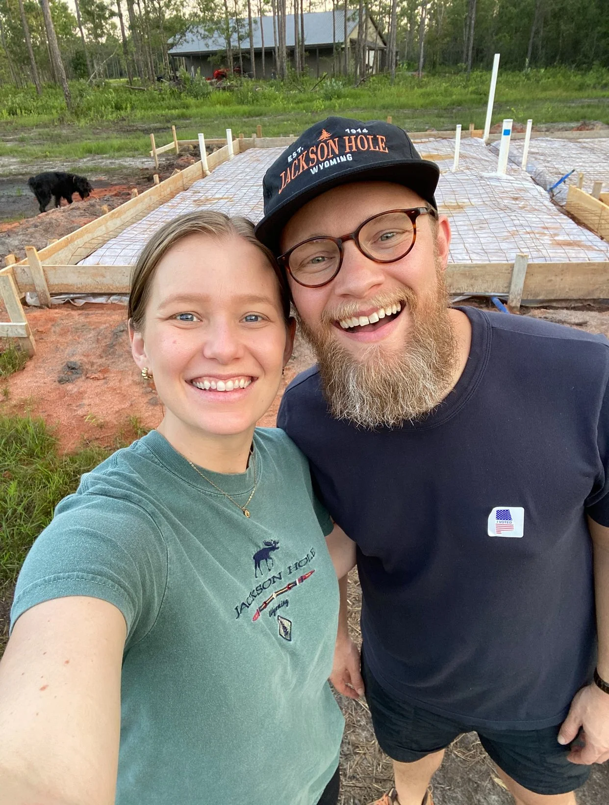 A smiling couple taking a selfie outdoors, with a construction site for a building foundation in the background, surrounded by trees.