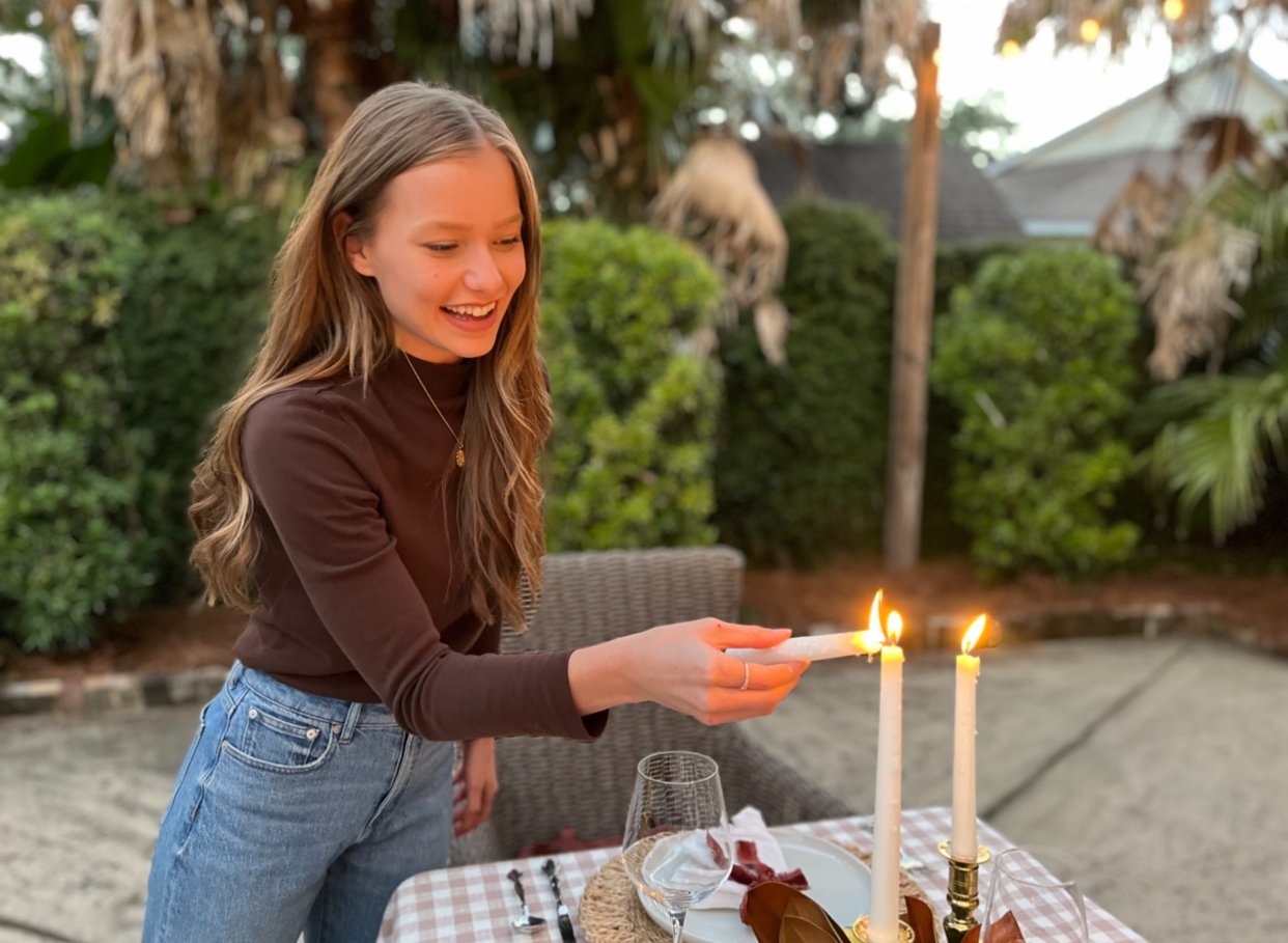 A young woman with long wavy hair, smiling and holding a lit candle, at an outdoor table set for a celebration with candles, plates, wine glass, and utensils, in a garden with greenery and palm trees.
