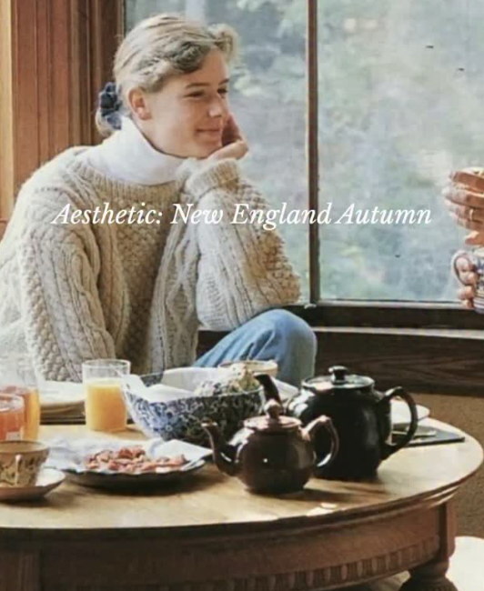 A woman with blonde hair in a bun, wearing a beige knitted sweater, sitting at a table by a window with a view of trees. She is resting her chin on her hand and looking out. The table has a teapot, cups, a glass of orange juice, and a plate of bacon.