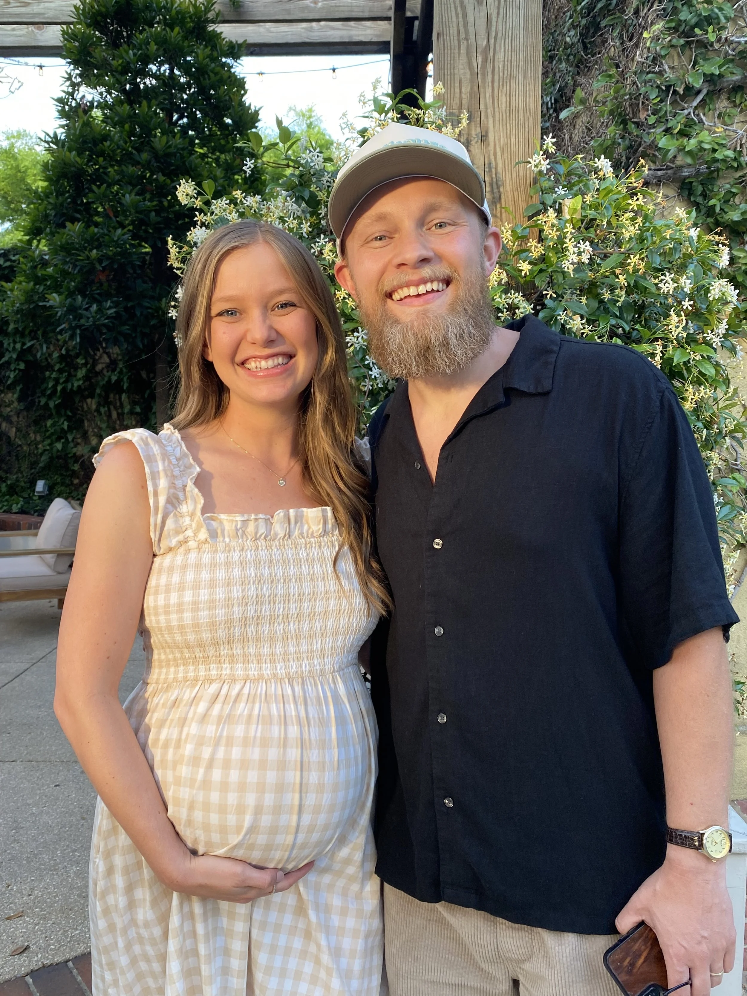 Pregnant woman in a checkered dress and a man in a black shirt smiling outdoors, standing in front of a green leafy background.