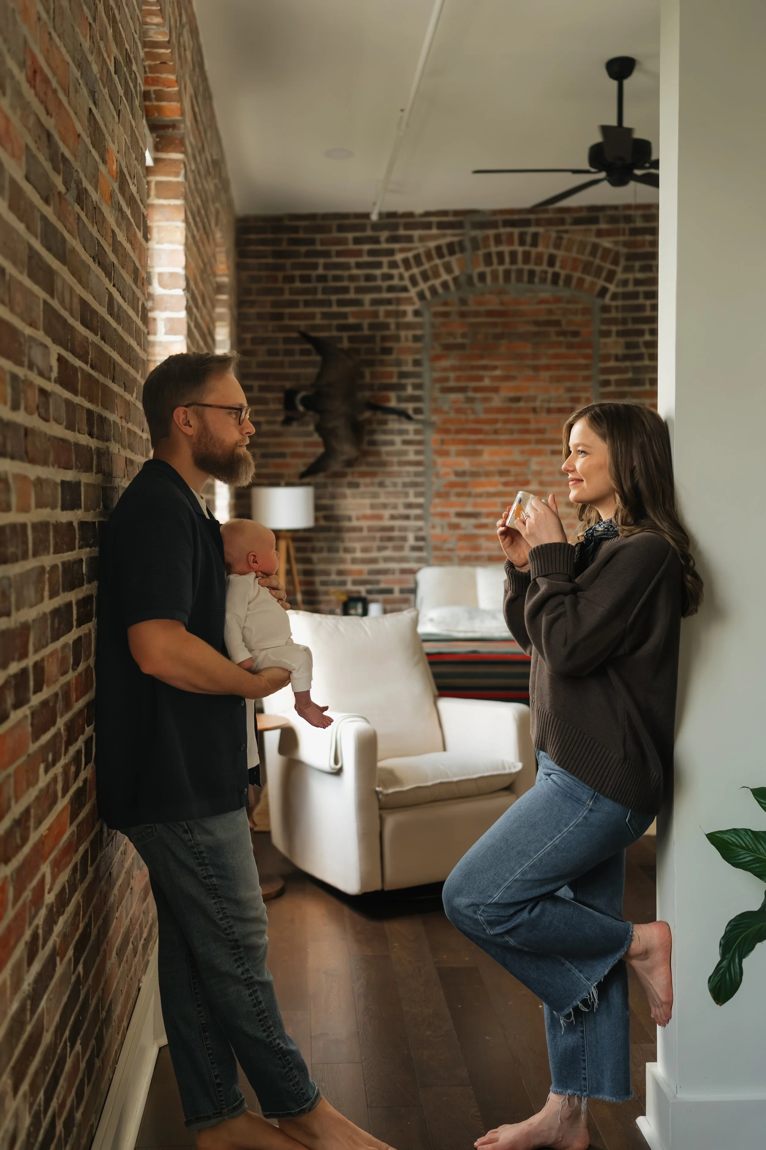 A young couple with a baby standing in a living room with a brick wall, smiling at each other. The woman is leaning against the wall, holding a coffee mug. The man is holding the baby in his arms. The room has a white armchair, a lamp, and a mounted 