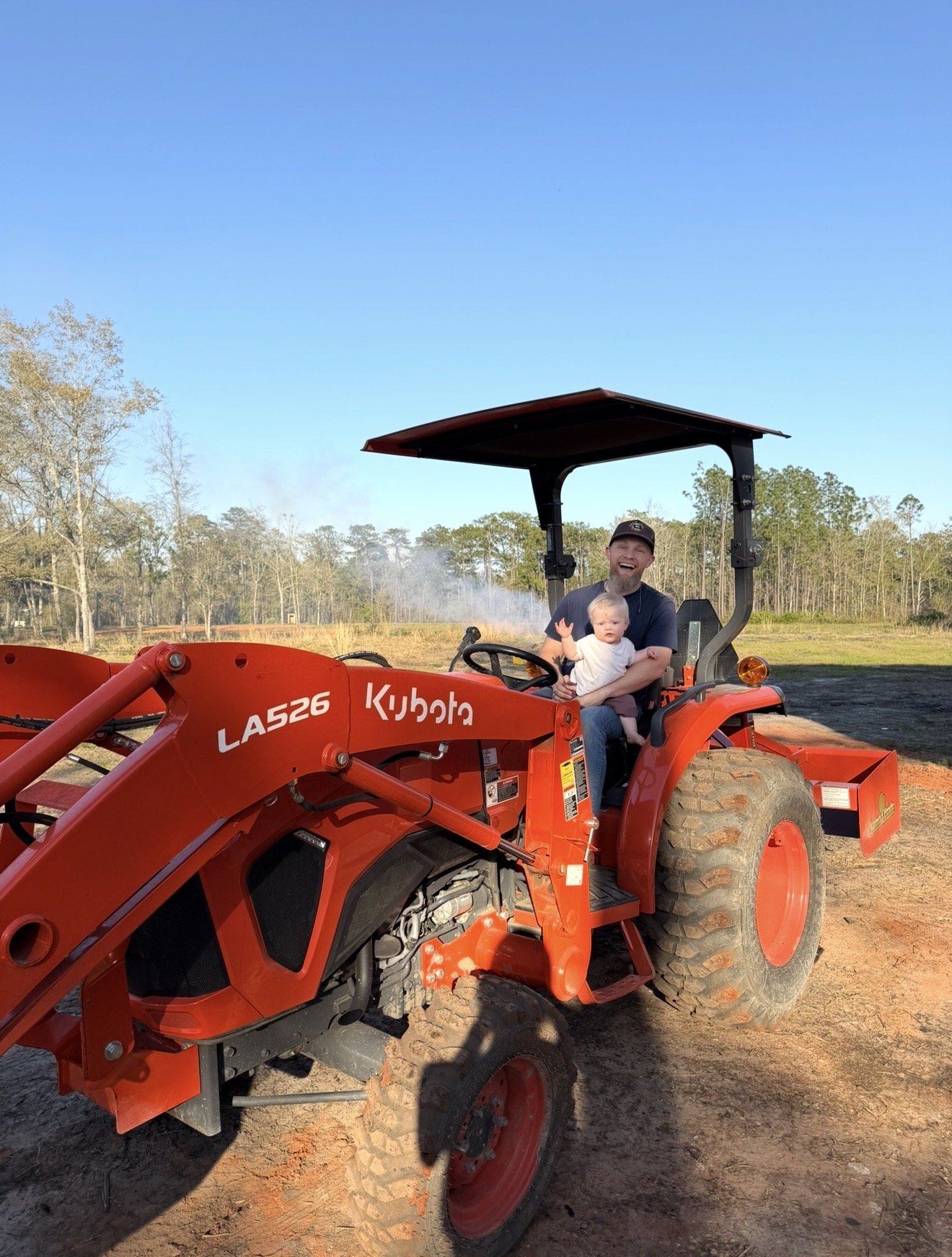 A man and a baby sitting on an orange Kubota tractor in a field with trees in the background under a clear blue sky.