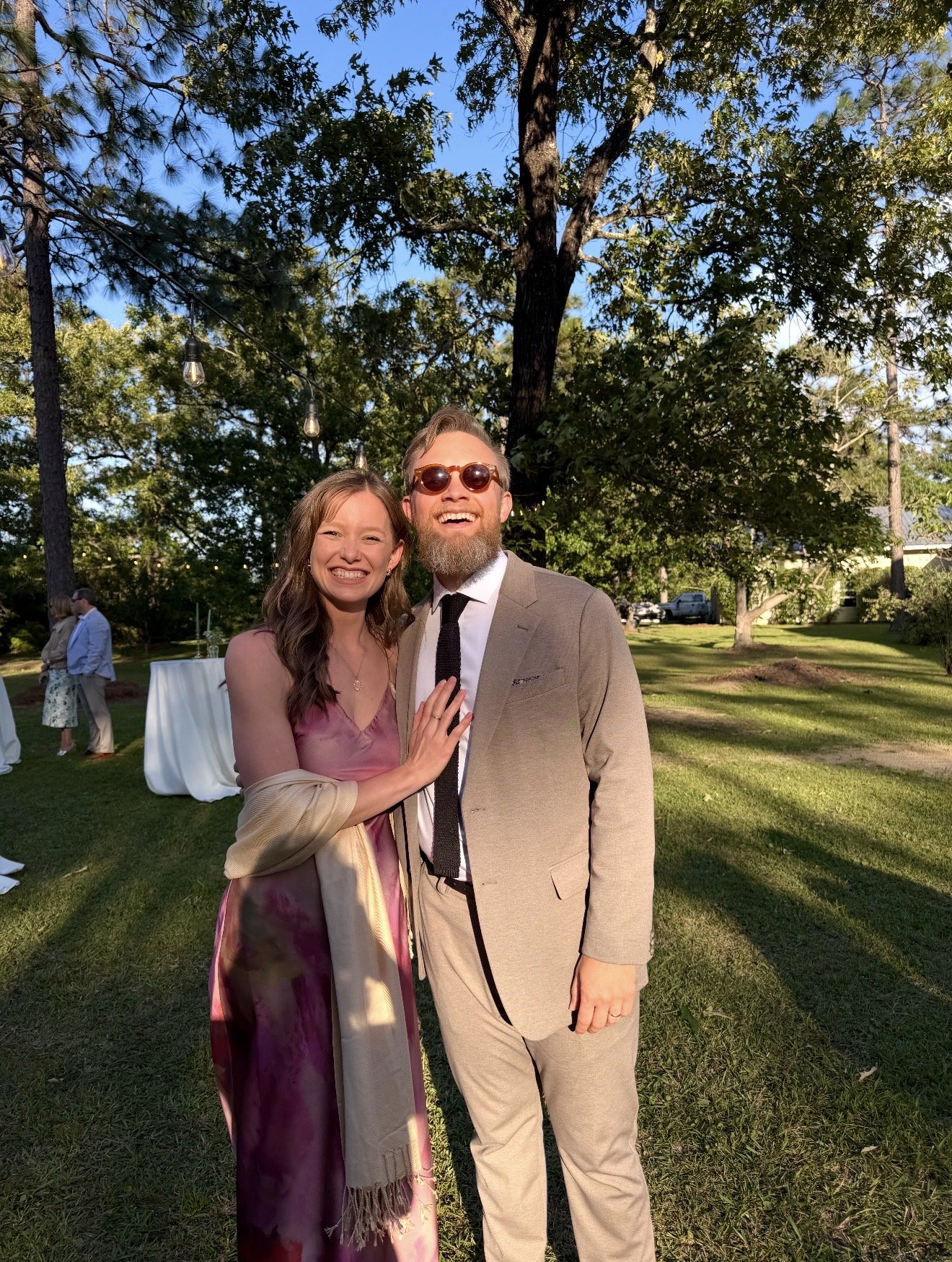 A woman and a man smiling and posing together outdoors during sunset in a grassy area with trees and string lights in the background. The woman is wearing a pinkish-purple dress and the man is dressed in a beige suit with sunglasses.