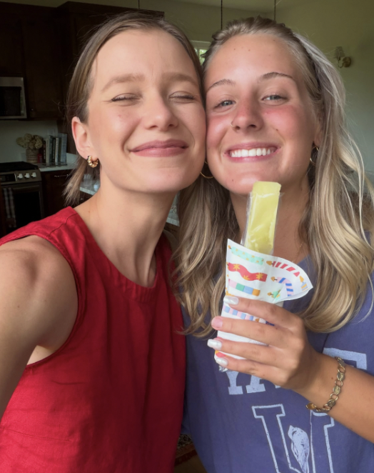 Two young women smiling and taking a selfie in a kitchen. One wears a red sleeveless top, and the other holds a colorful paper craft with a popsicle.