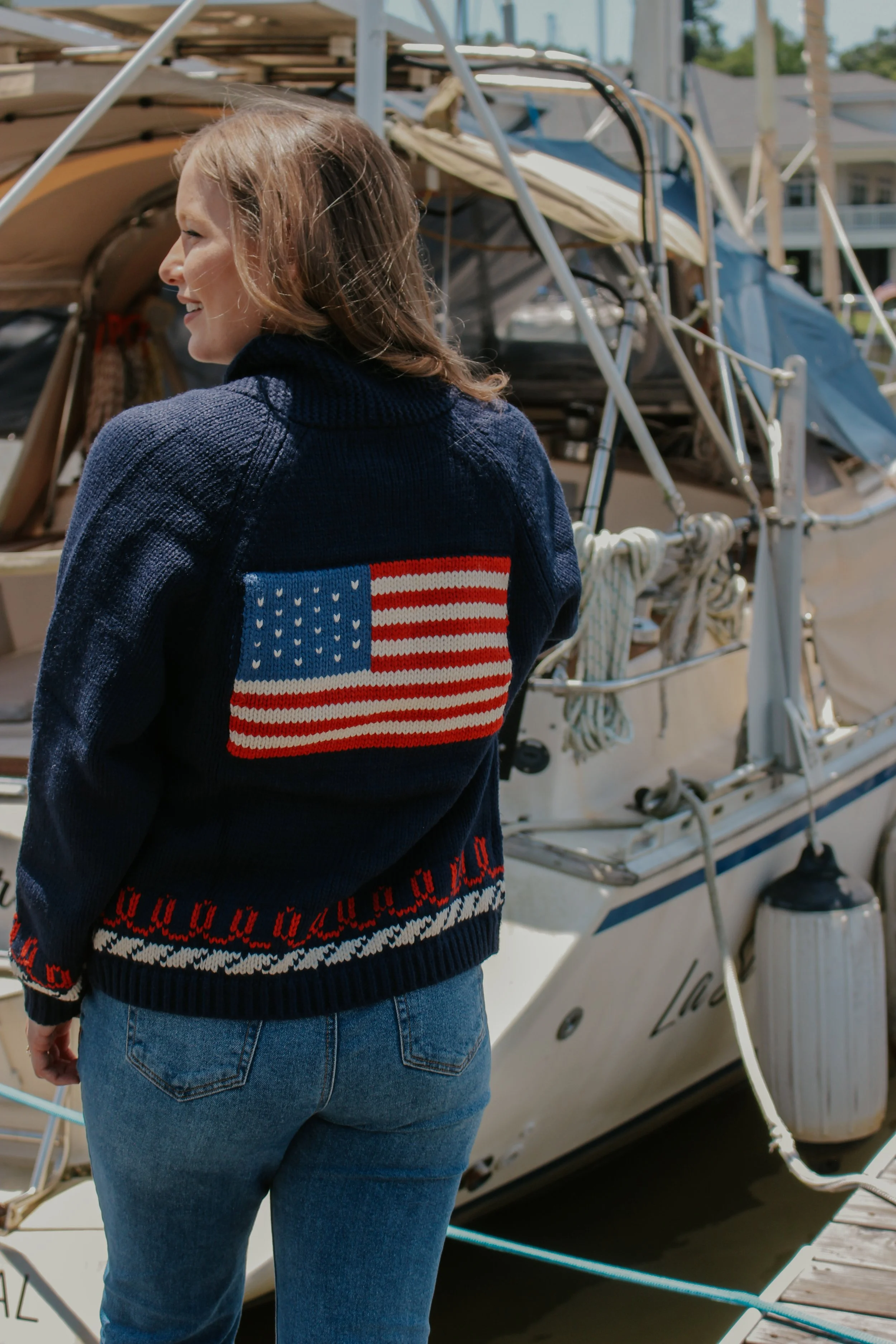 Woman wearing a navy sweater with an American flag patch on the back, standing near a sailboat docked at a marina.