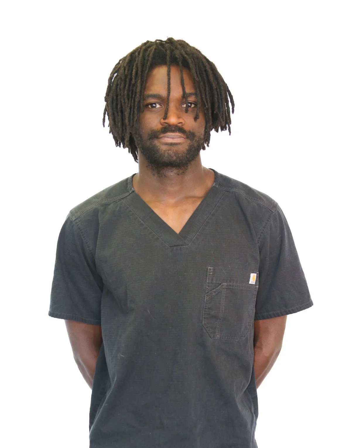 Portrait of a man with dark skin, curly black hair, and a beard, wearing black medical scrubs, standing against a plain white background.