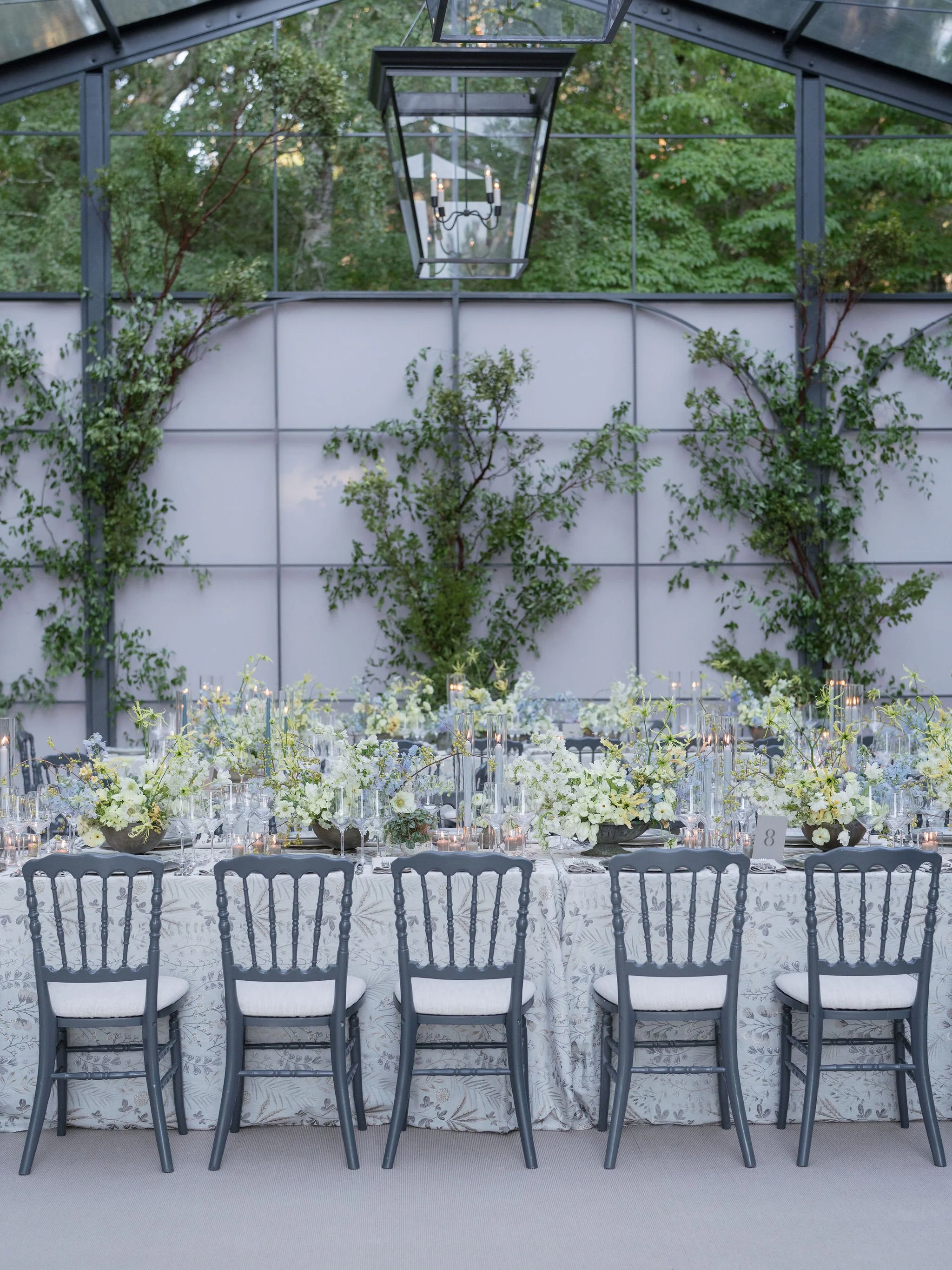 A long banquet table decorated with white flowers, candles, and glassware, with a backdrop of a wall adorned with green climbing plants.