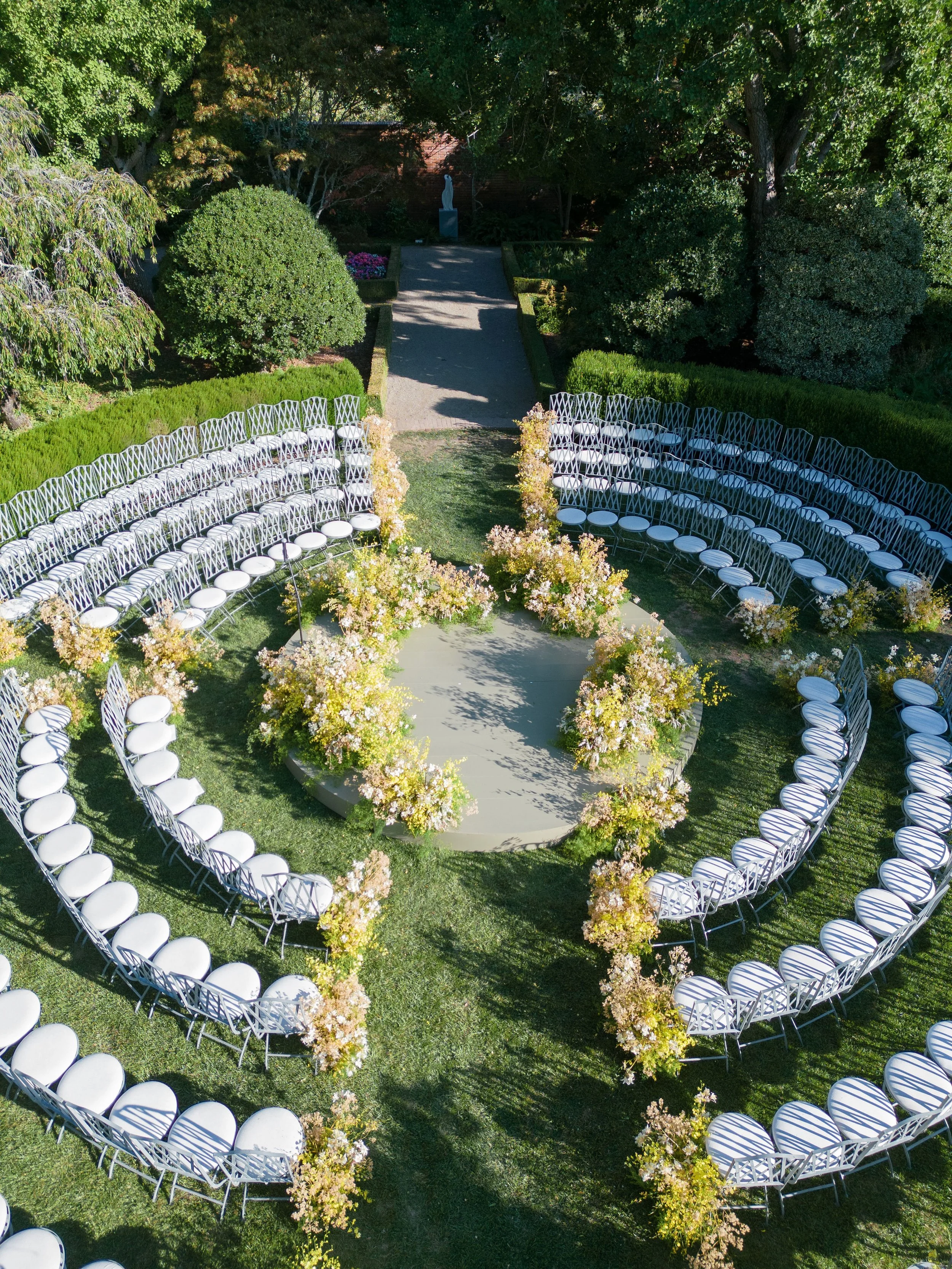 Outdoor wedding round ceremony setup with white chairs arranged in a heart shape and a floral aisle leading to a statue and a backdrop of lush green trees.