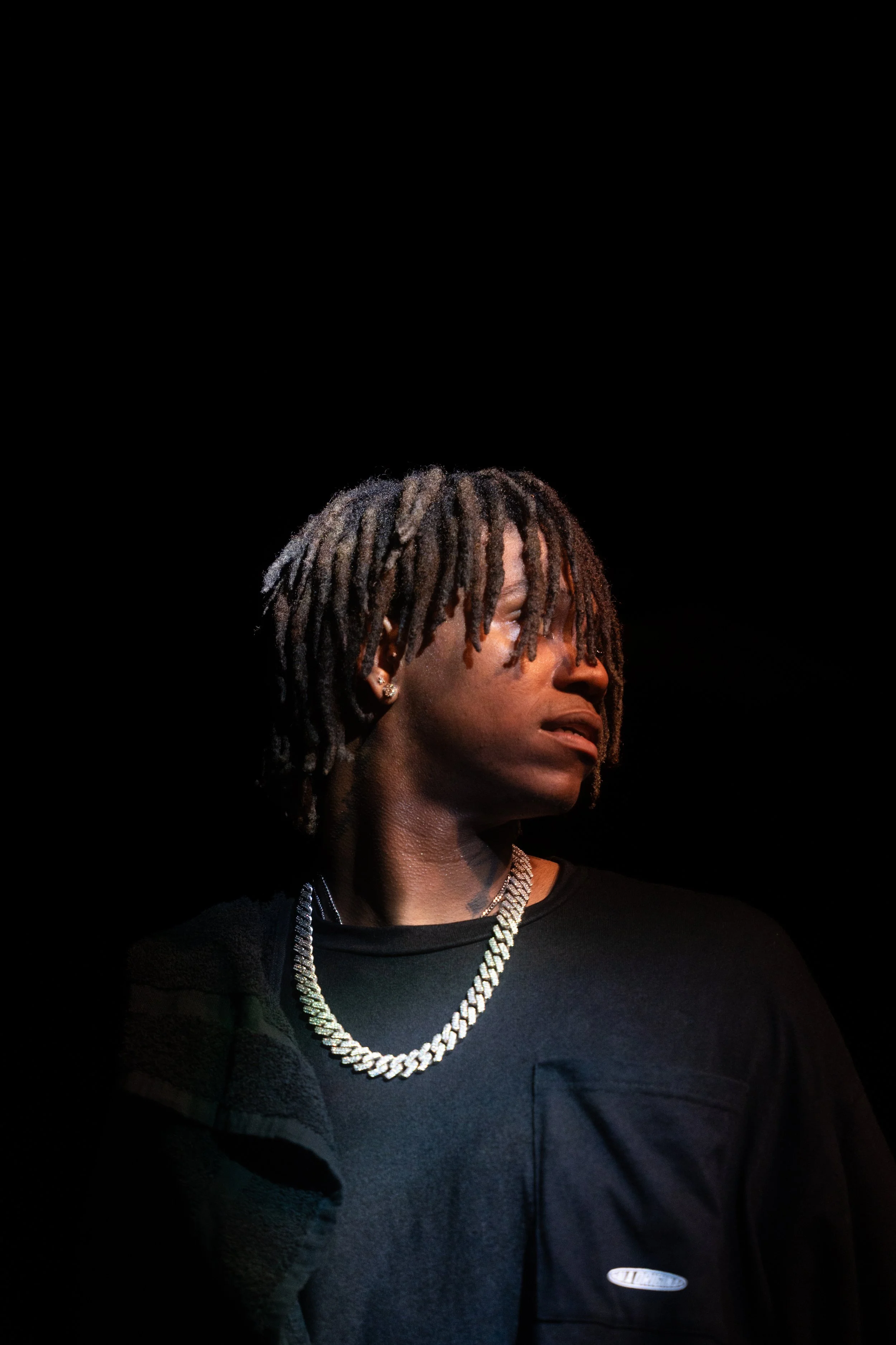 Person with dreadlocks wearing a silver chain and dark shirt against a black background.