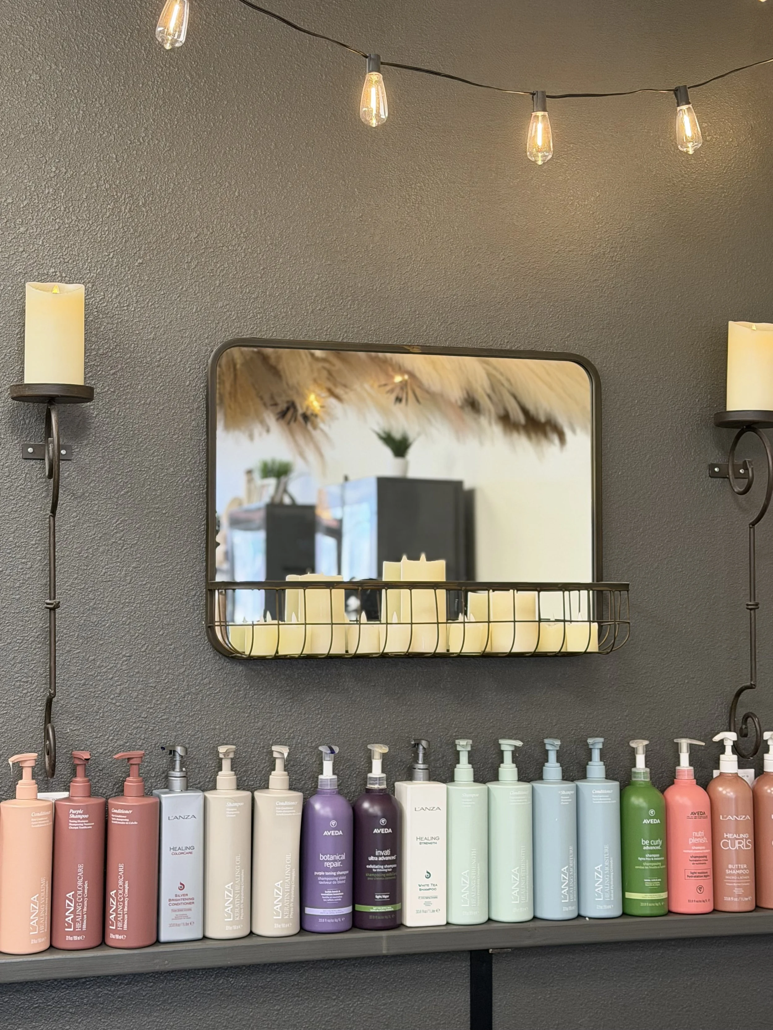 A wall with string lights and wall-mounted candle holders, a mirror with candles on its shelf, and a row of colorful bottles of hair care products below.