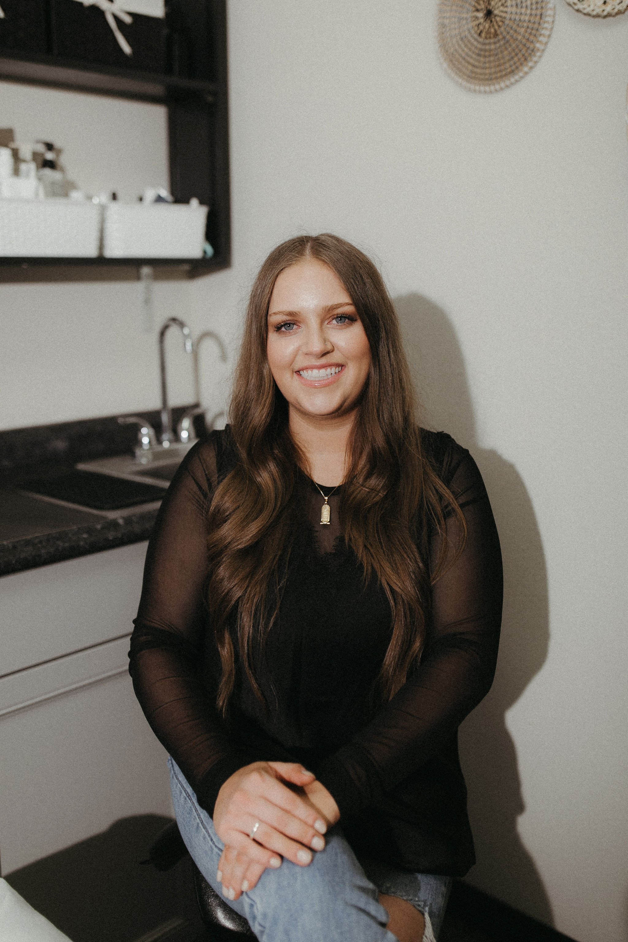 A smiling woman with long brown hair, wearing a black sheer top and denim jeans, sitting with her hands clasped in her lap in a room with a kitchen in the background.