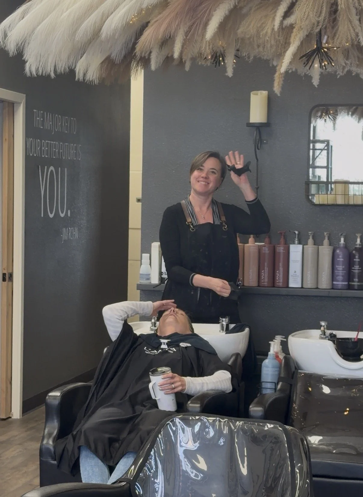 A woman getting her hair washed at a salon, smiling and waving, while a stylist stands behind her. The salon has a dark gray wall with a quote, shelves with hair products, and hair washing stations.