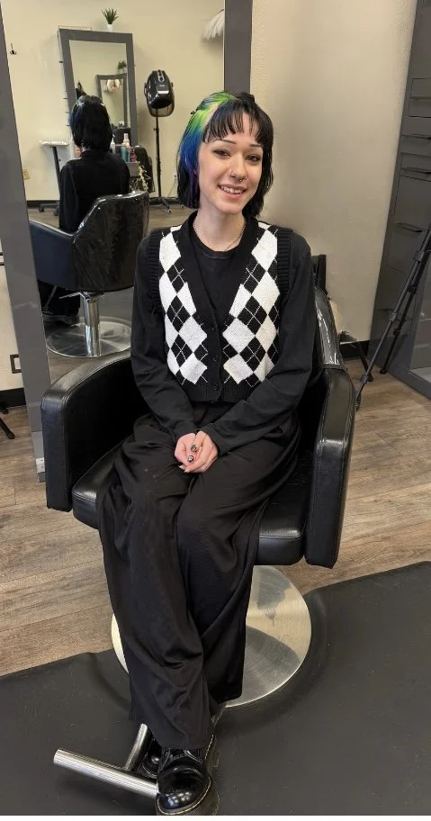 Young woman with multi-colored hair, sitting in a salon chair, smiling, wearing a black and white argyle vest over a black outfit, with a mirror and salon equipment visible in the background.