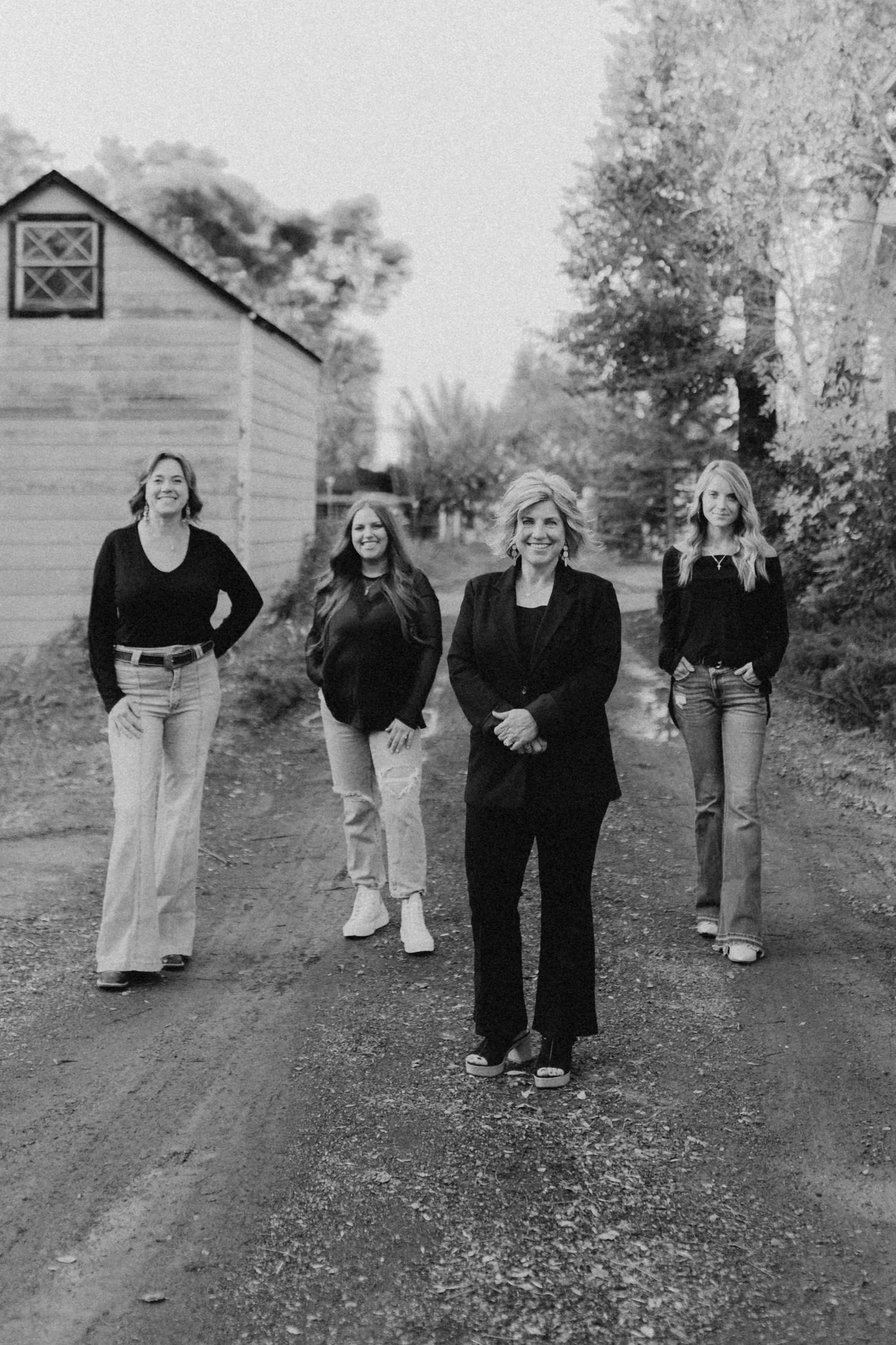 Black and white photo of four women walking down a dirt road, smiling, with trees and a barn in the background.