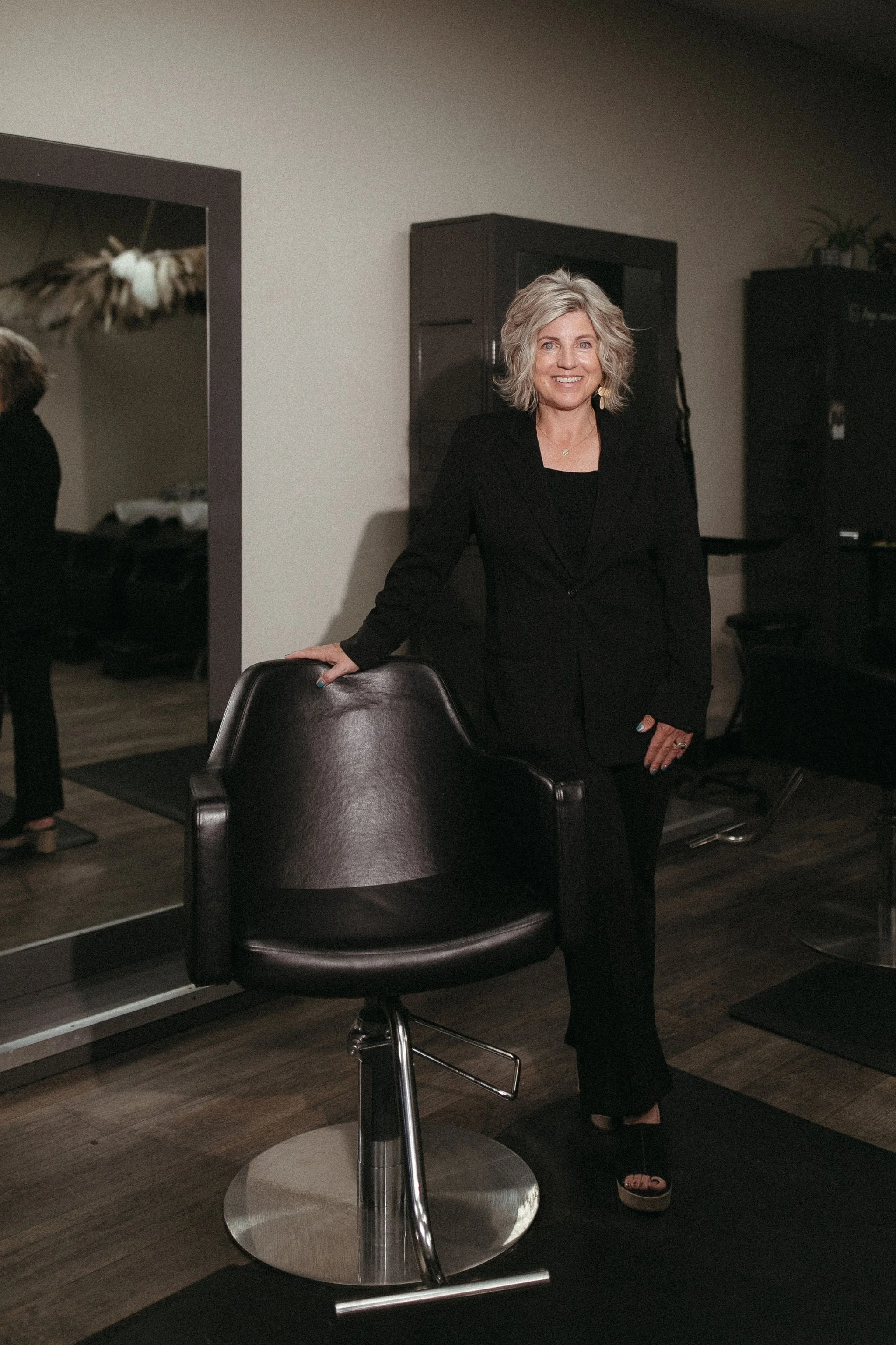 A woman with curly gray hair in a black blazer and pants, standing next to a black salon chair in a hair salon or studio.