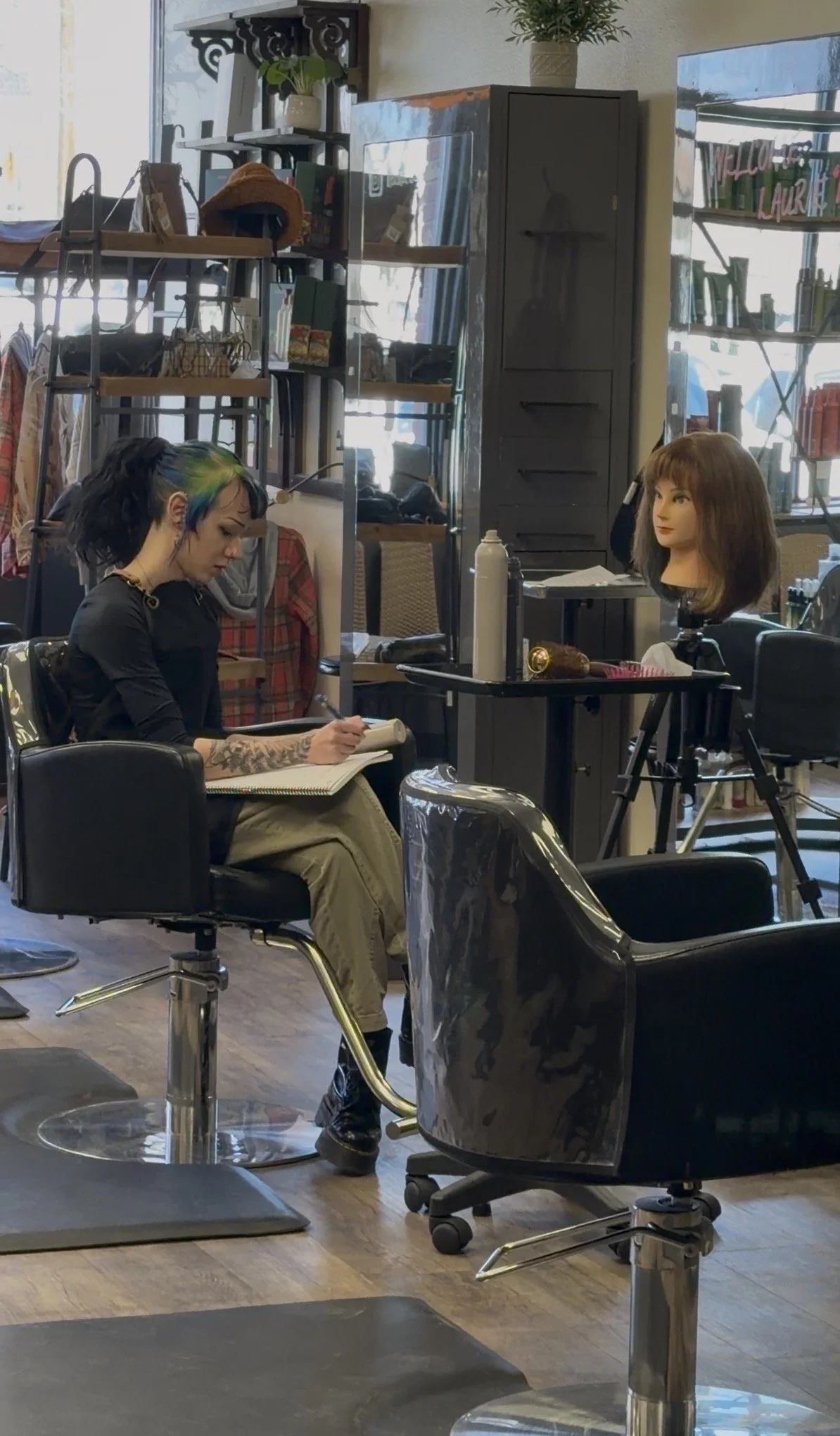 A person with colorful hair taking notes in a hair salon with mannequin heads and styling tools.