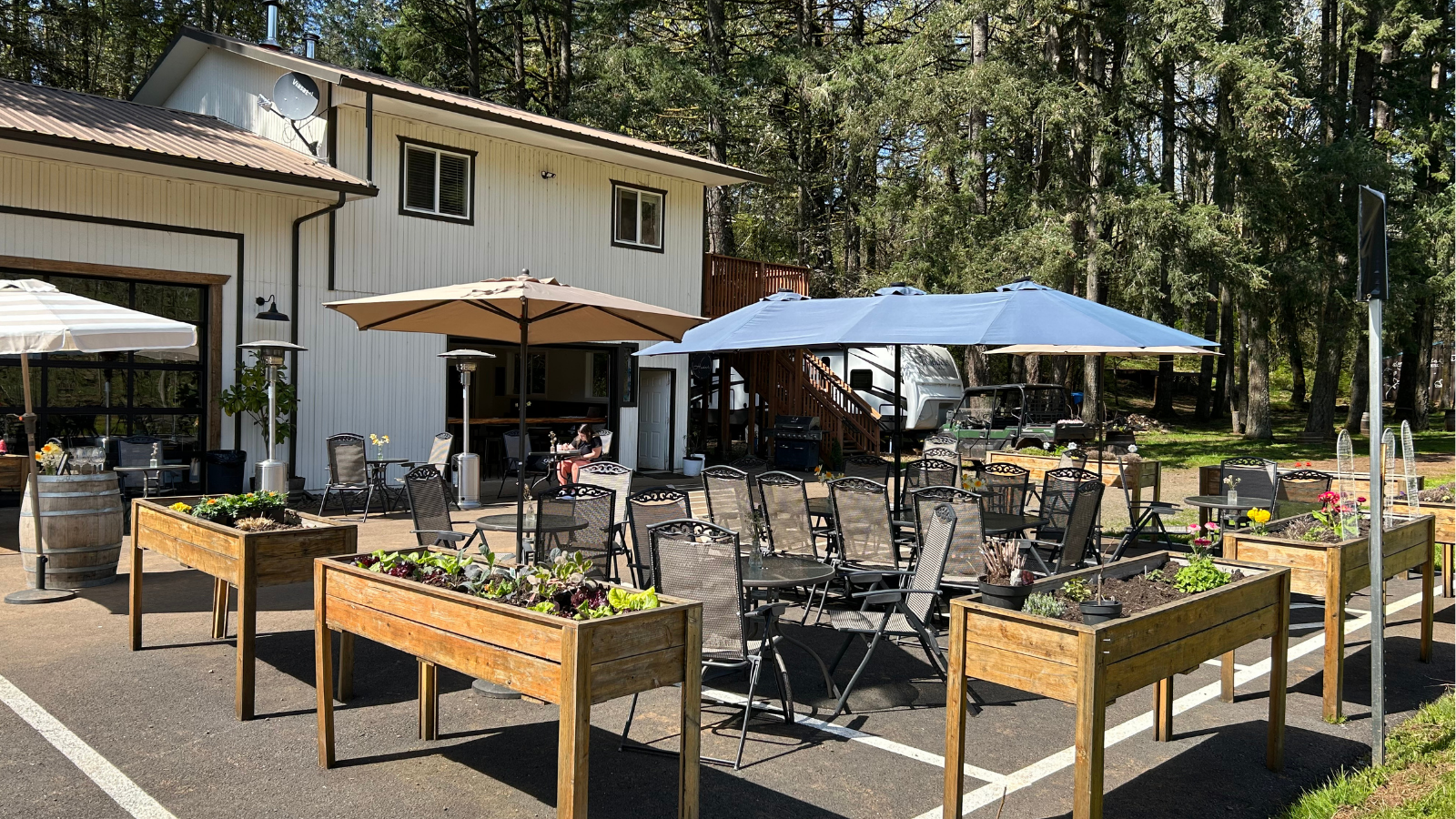 An outdoor patio area with tables, chairs, and umbrellas in front of a white building with stairs leading to an upper level, surrounded by trees. There are planters with flowers and a person sitting at a table.