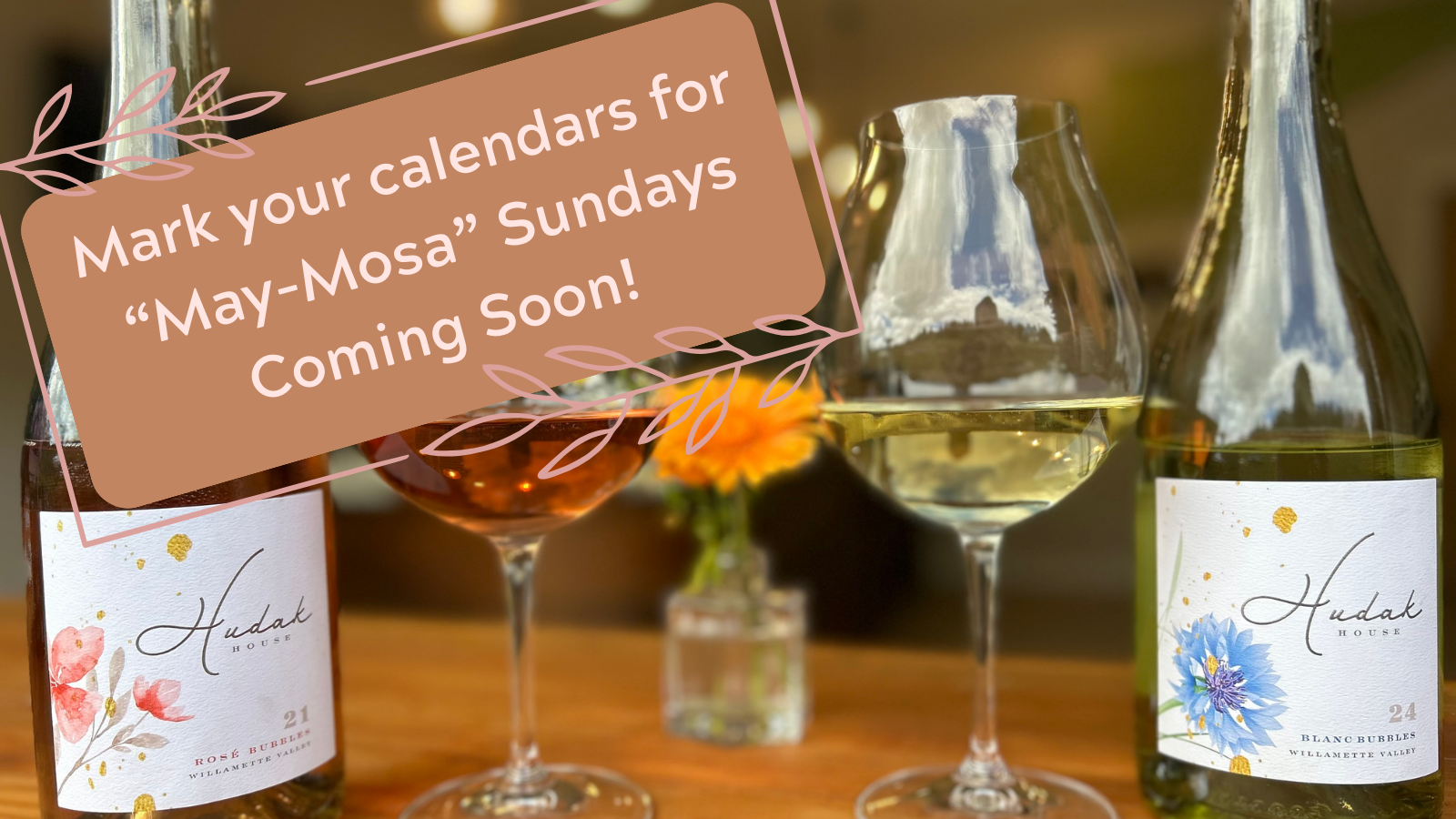Different glasses of wine on a table with a small flower vase in the background, with a sign indicating upcoming 'May-Mosa' Sundays at Hudak House.